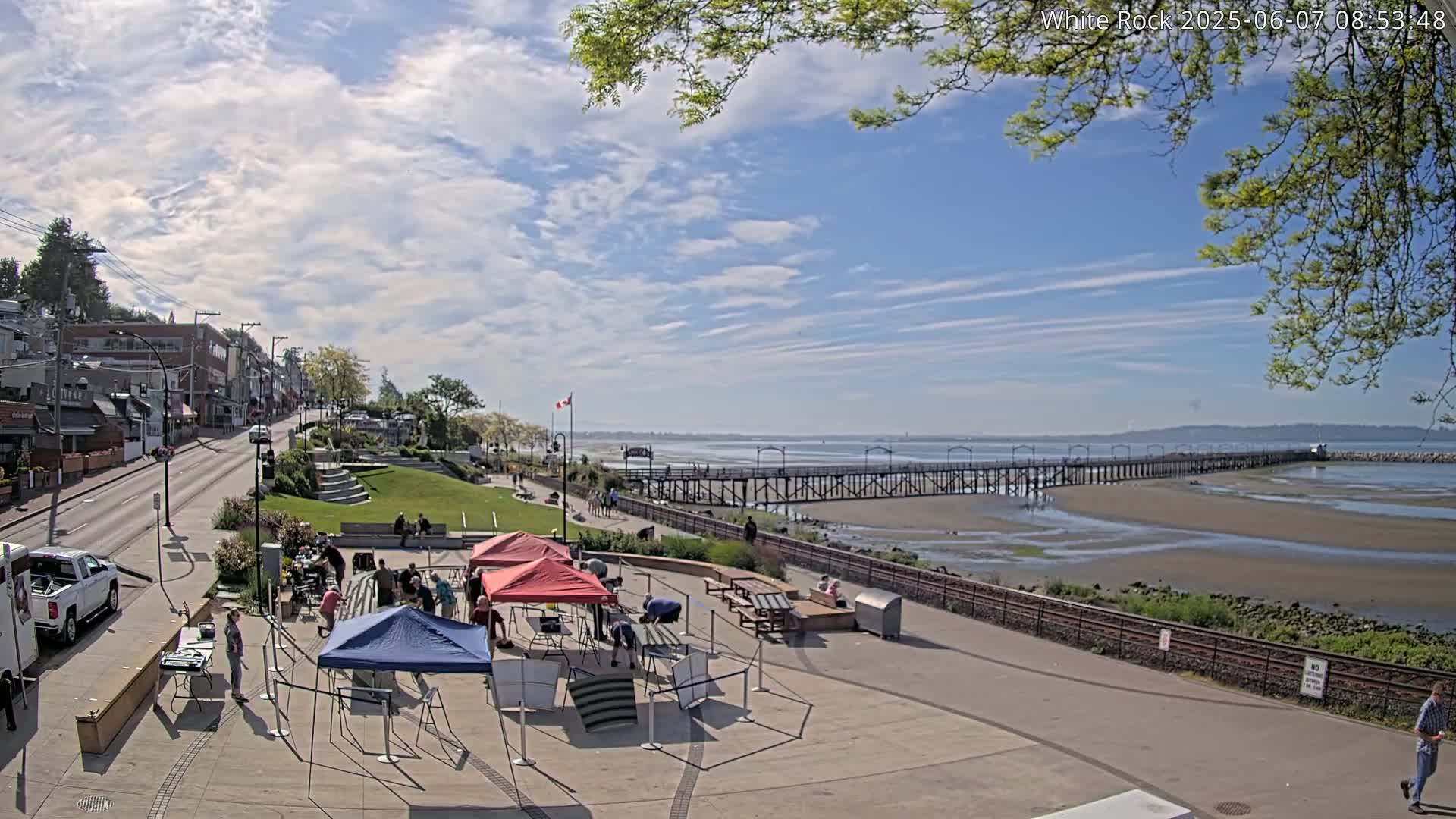 A seaside town on a partly sunny day with a long pier extending into a tidal flat, features several people gathered near tents in a plaza area next to a street lined with buildings.