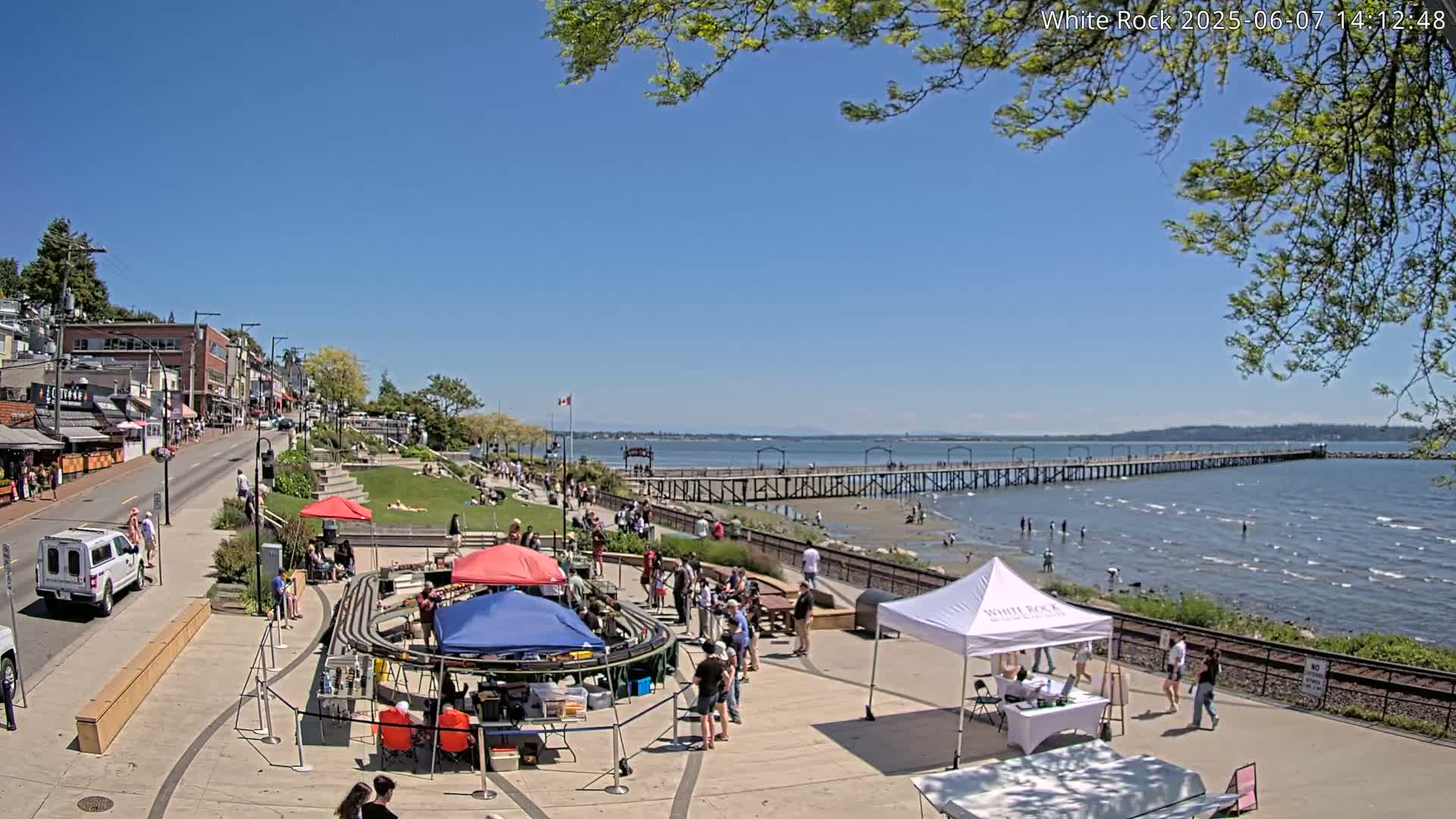 A sunny seaside town scene shows a long pier extending into a calm body of water, with people gathered on a paved area near shops and a grassy park, under a mostly clear blue sky.