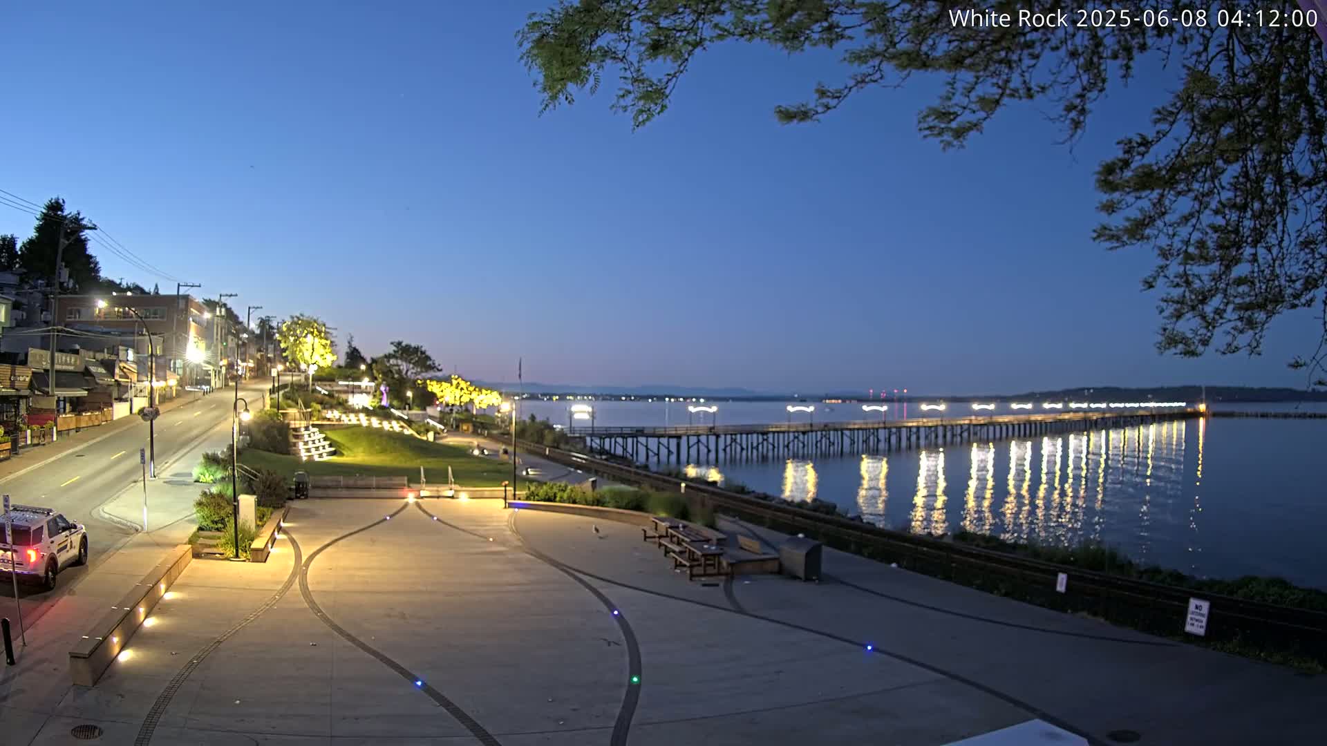 A long pier stretches across calm water at twilight, reflecting lights onto the water's surface, next to a paved waterfront area with benches and a street lined with buildings.