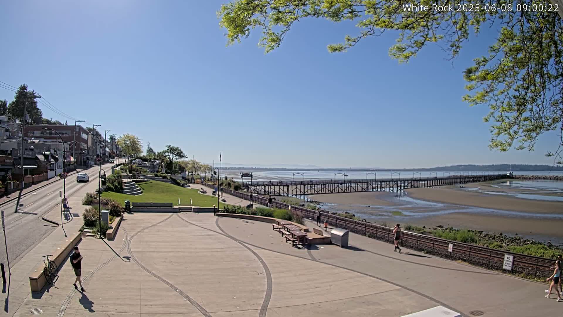 A paved waterfront plaza overlooks a long pier extending into a calm, sunny bay, with a few people walking along the shore and a town visible in the background.