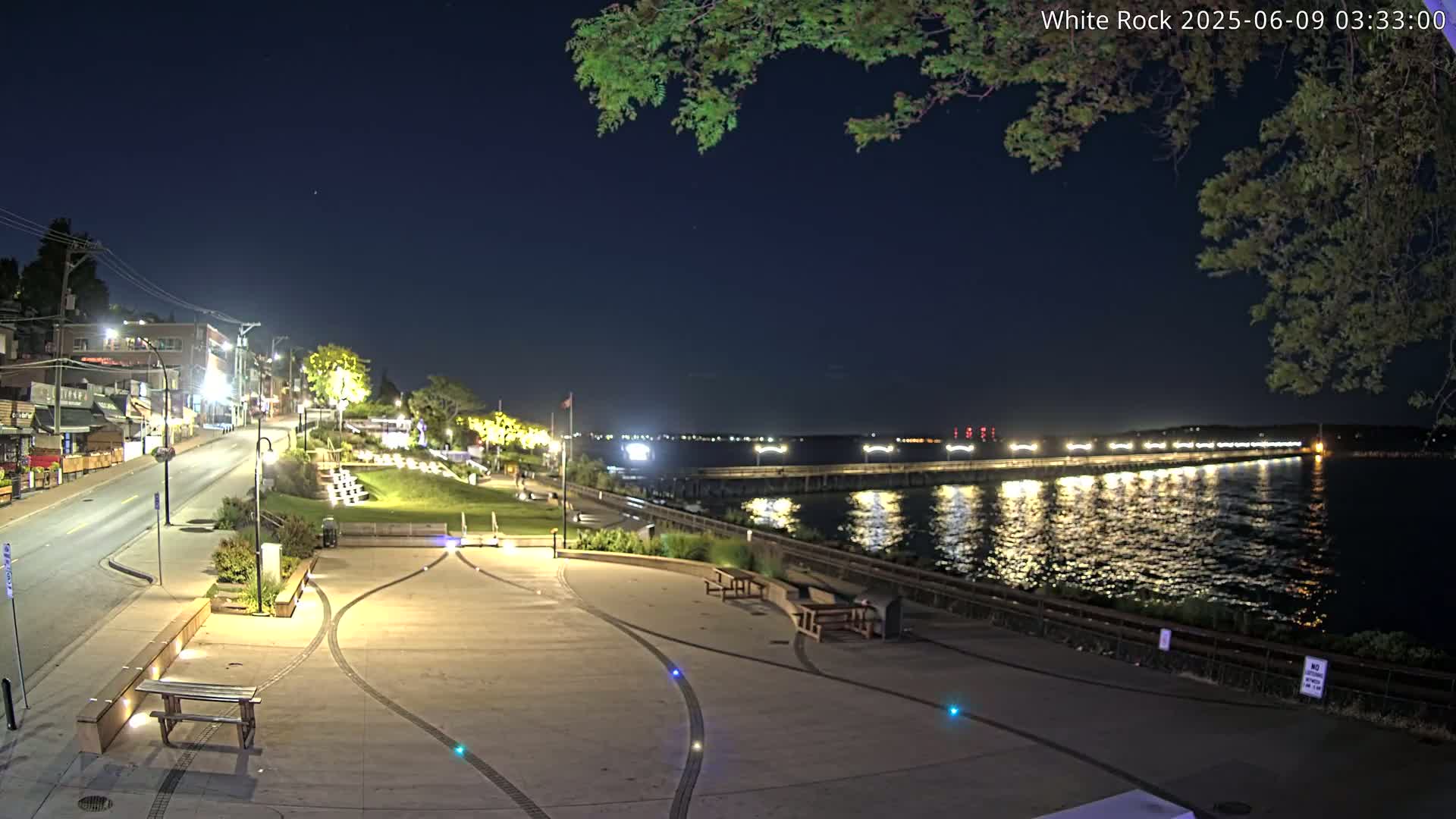 A nighttime view of a waterfront town with a paved walkway, benches, and a pier extending into the calm water under a clear, dark sky.