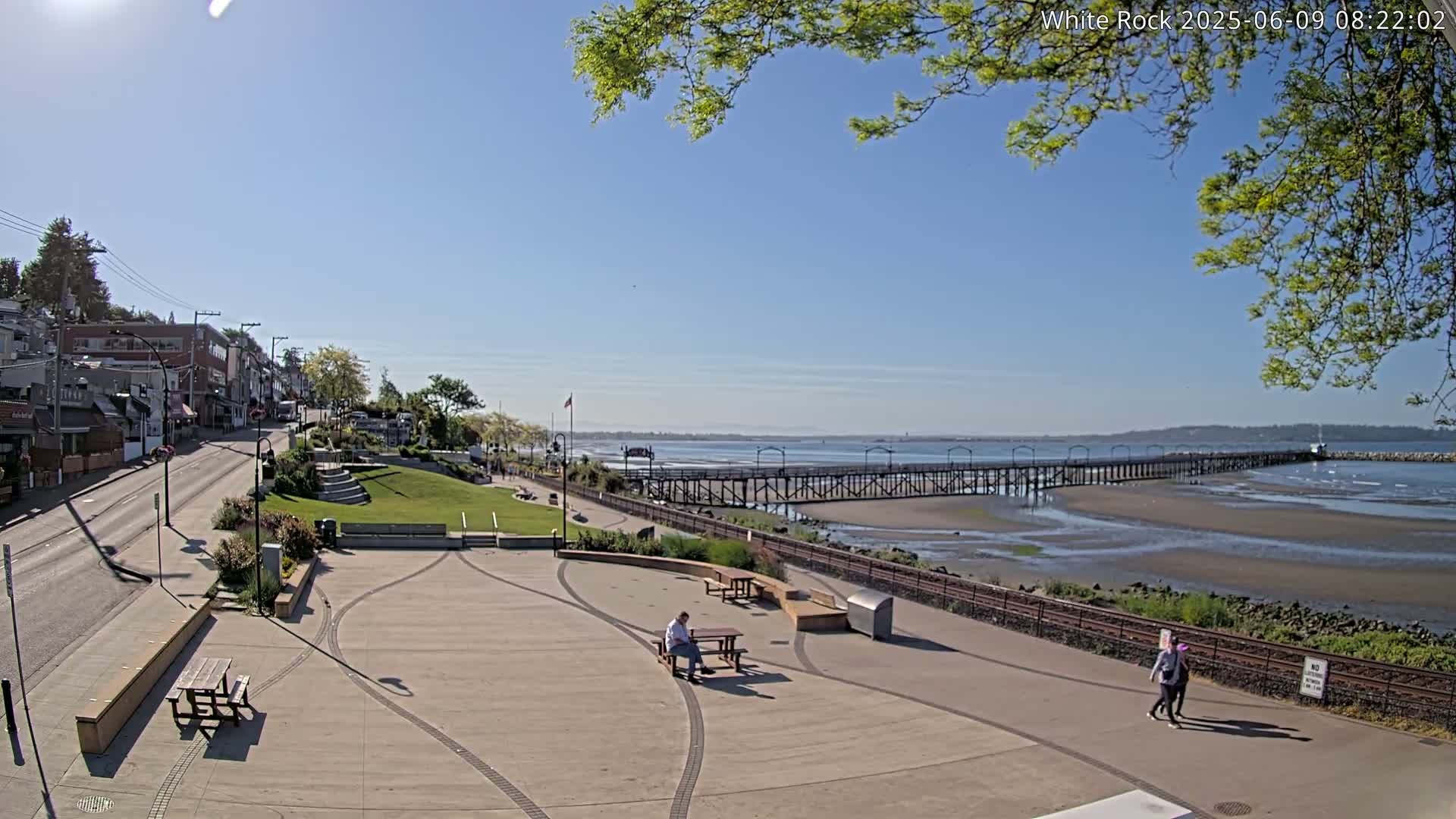 A paved waterfront plaza with picnic tables and a few people, overlooks a long pier extending into a calm bay under a clear, sunny sky.