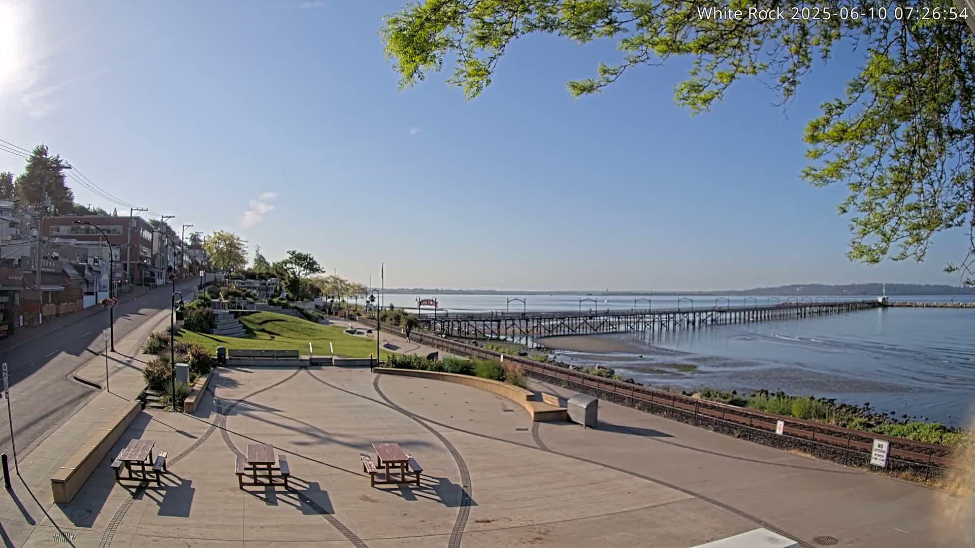 A paved waterfront park with picnic tables overlooks a long pier extending into a calm bay under a sunny sky.