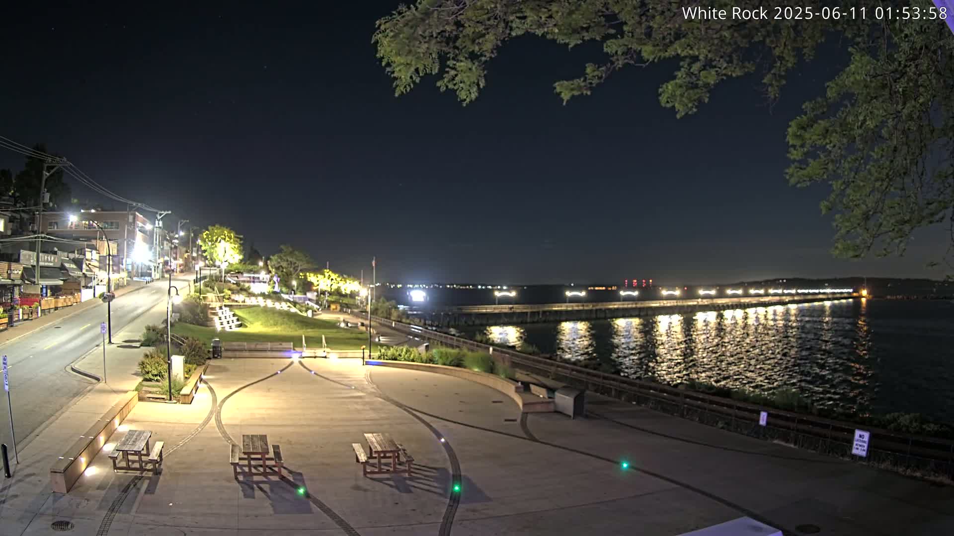 A nighttime view of a waterfront park with picnic tables, a paved walkway, and a long pier extending into a calm body of water under a clear, dark sky.