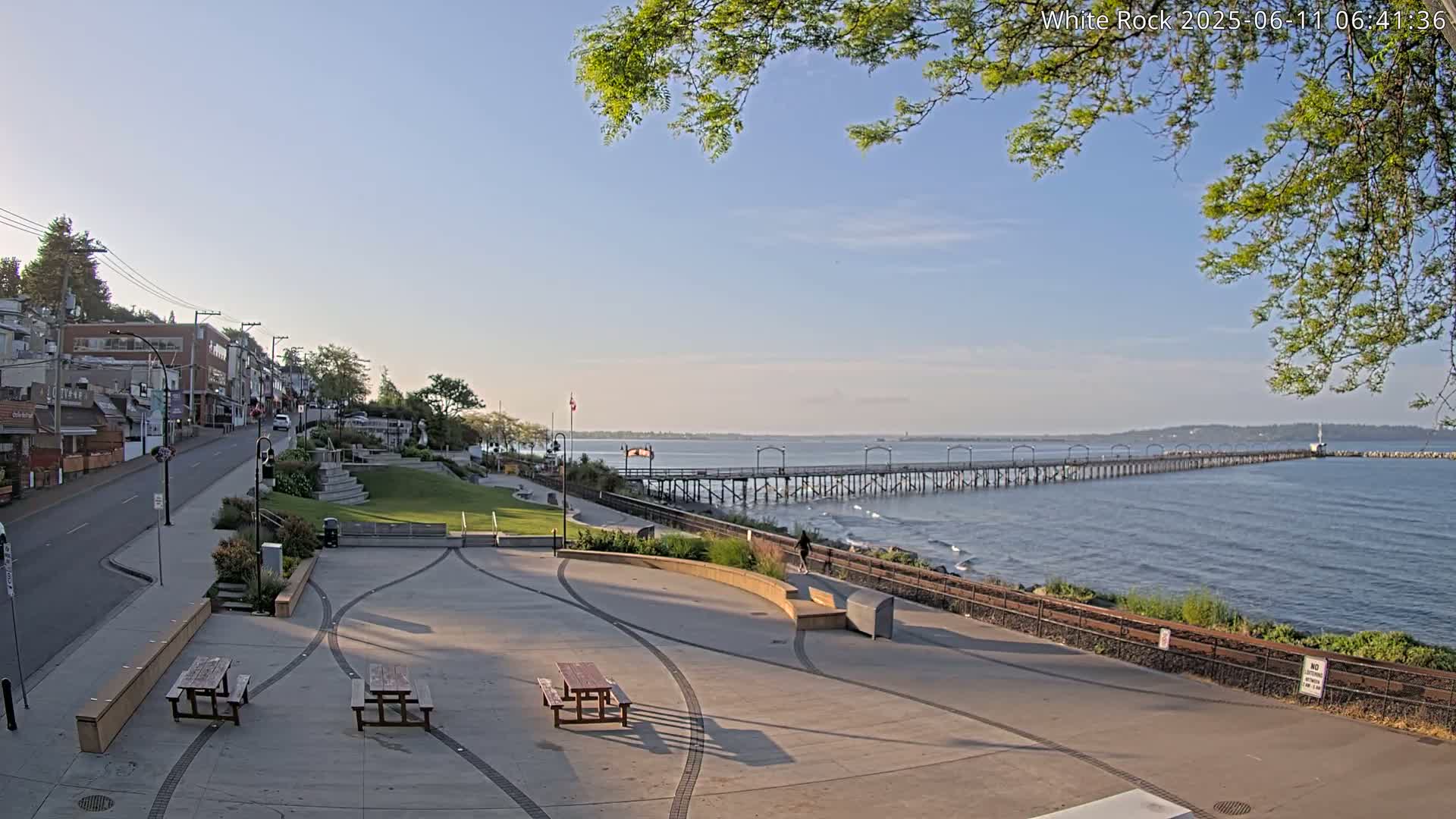 A long pier extends into a calm ocean under a mostly clear, sunny sky, overlooking a paved waterfront plaza with picnic tables and a street lined with buildings.