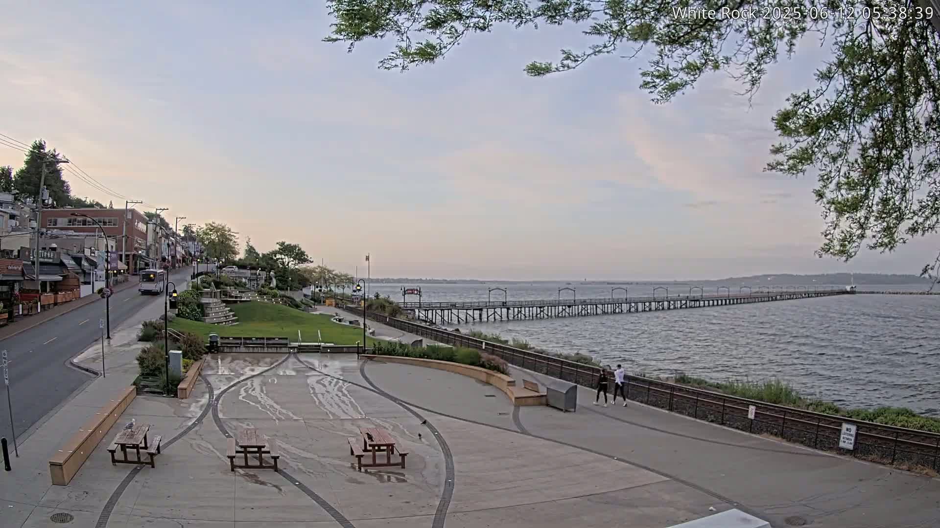 A paved waterfront area with picnic tables and a long pier extending into a calm body of water under an overcast sky.