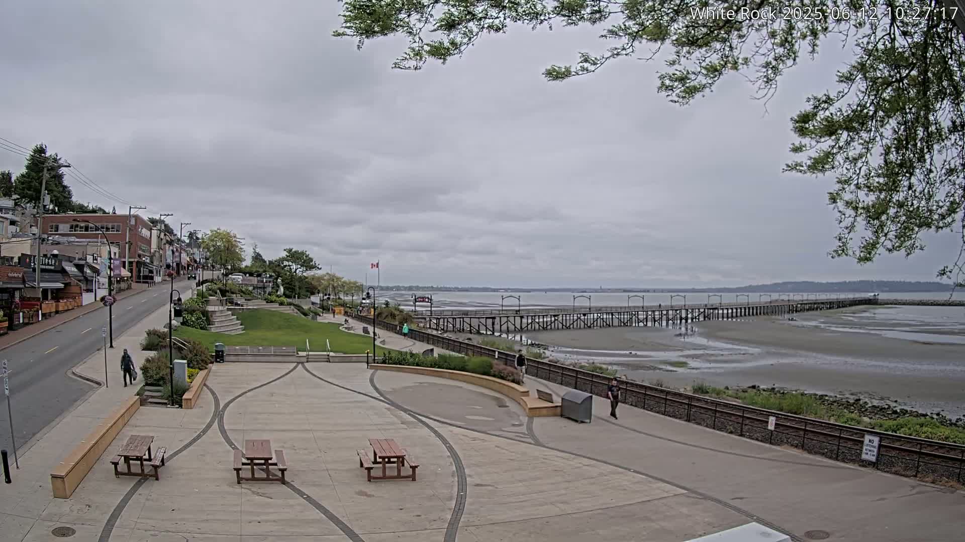 A paved plaza with picnic tables overlooks a long pier extending into a calm, grey-sky day at a beach with exposed mudflats.