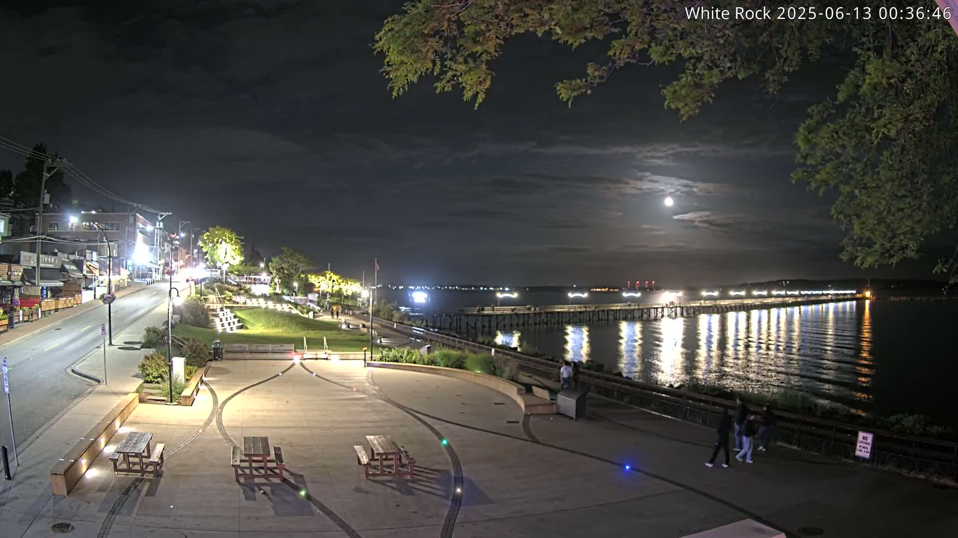 A nighttime view of a waterfront town with a long pier extending into a calm, moonlit body of water, featuring a paved plaza with picnic tables and a street leading to buildings beyond.