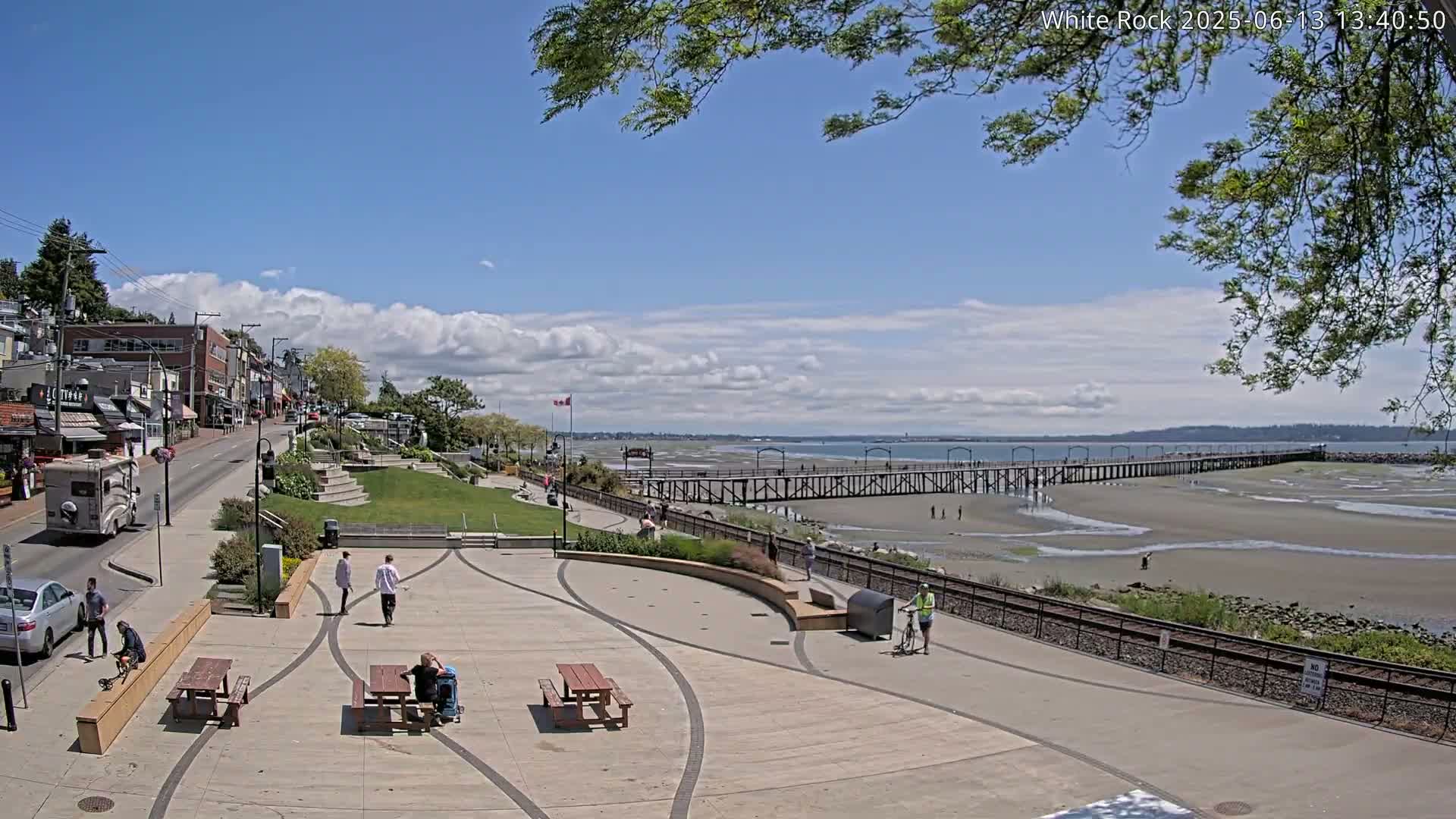A partly cloudy day shows a seaside town with a long pier extending into the exposed sand flats, a paved plaza with picnic tables and a few people walking around.