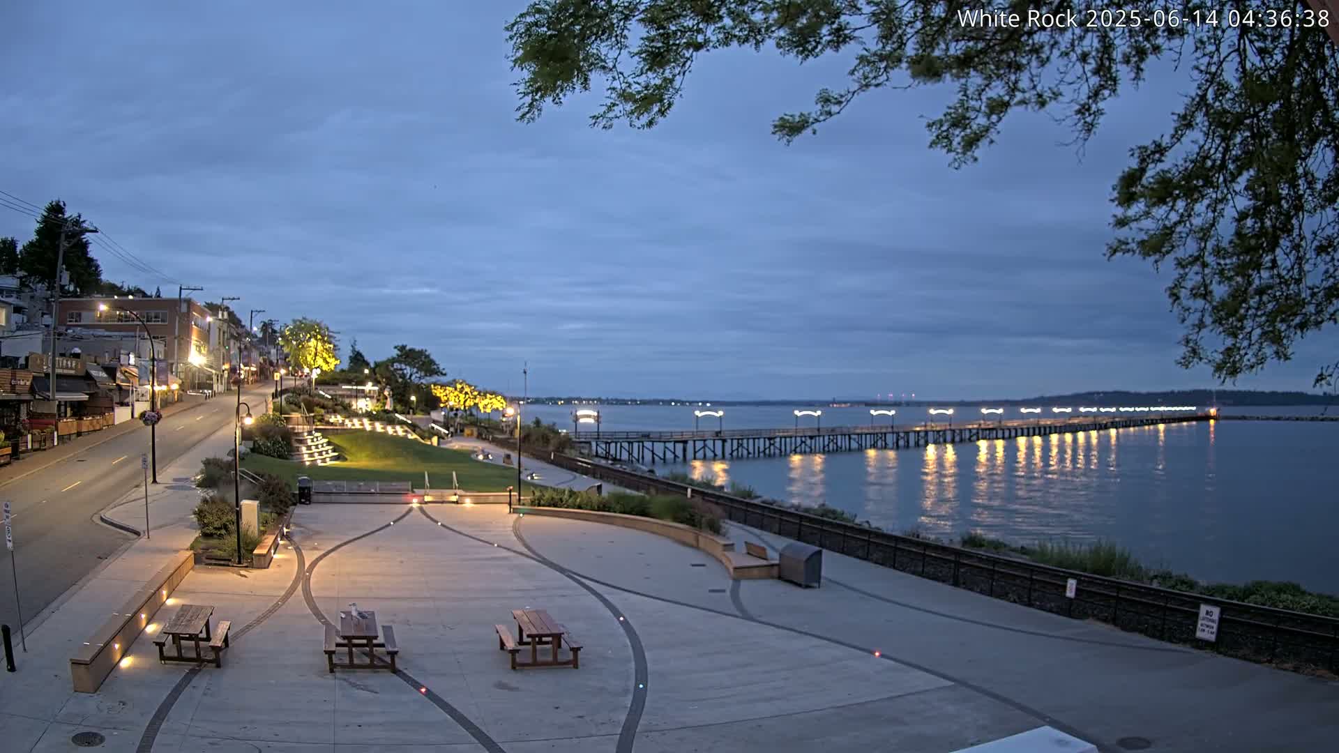 A paved waterfront area with picnic tables overlooks a long, lit pier extending into a calm, twilight sea.