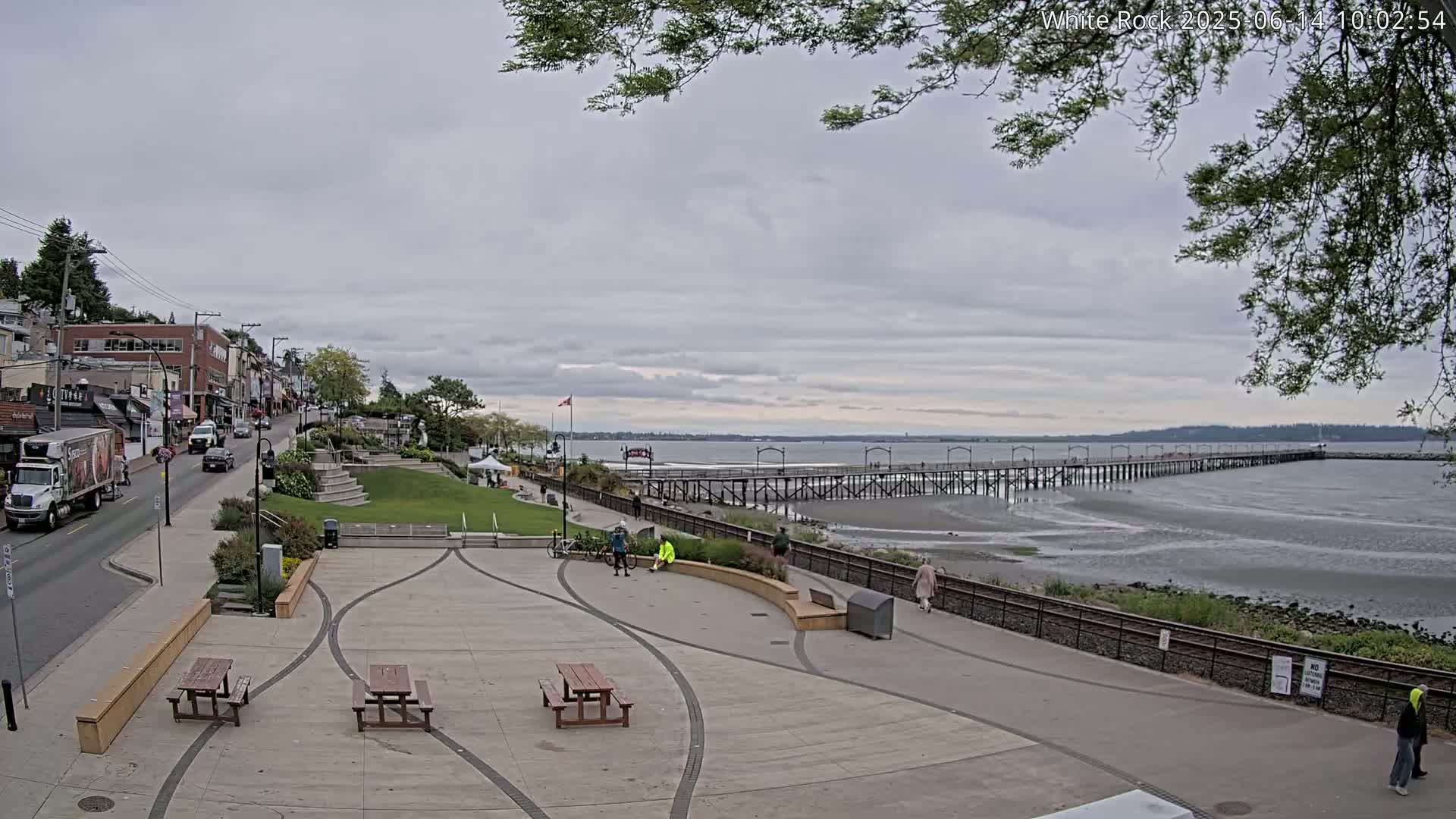 A paved plaza with picnic tables overlooks a long pier extending into a calm, gray-sky day, with a street and buildings visible in the background.