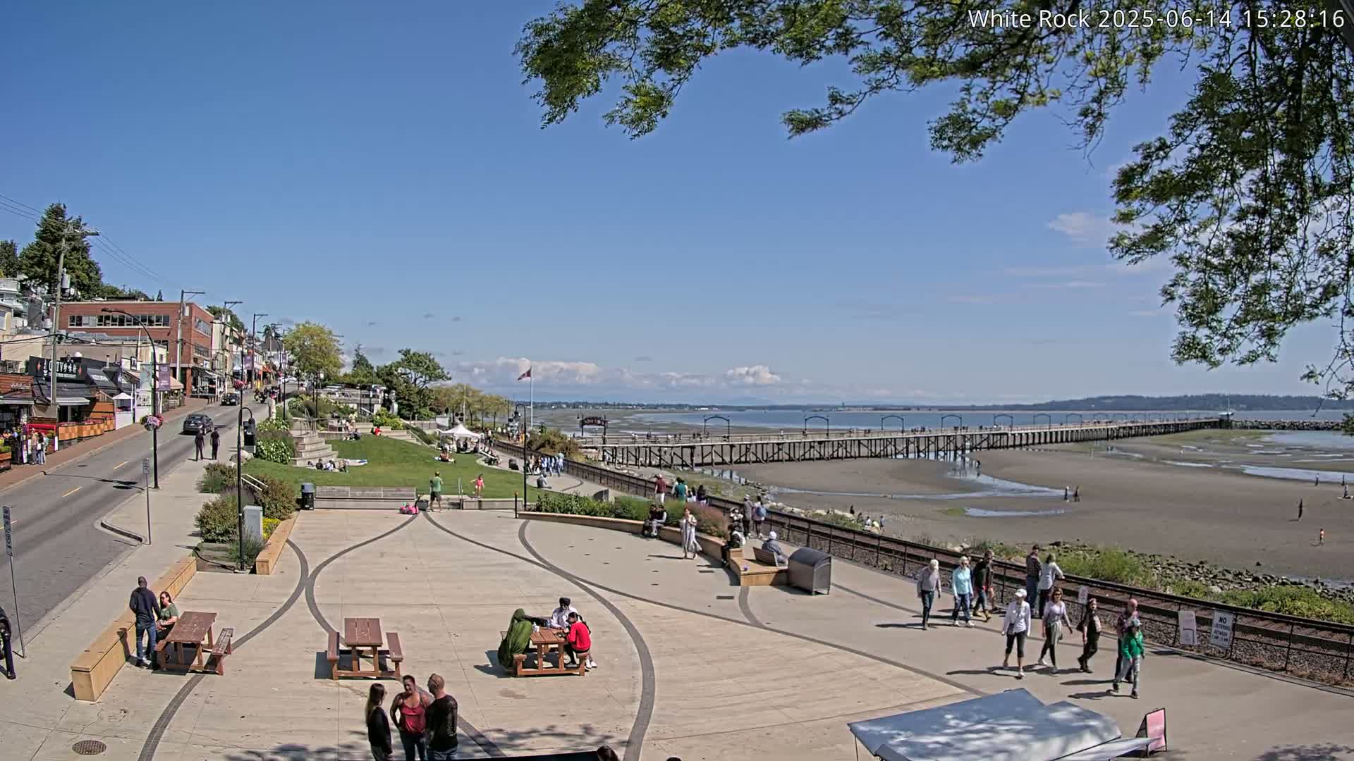 A paved plaza with picnic tables overlooks a long pier extending into a calm, gray-sky day, with a street and buildings visible in the background.