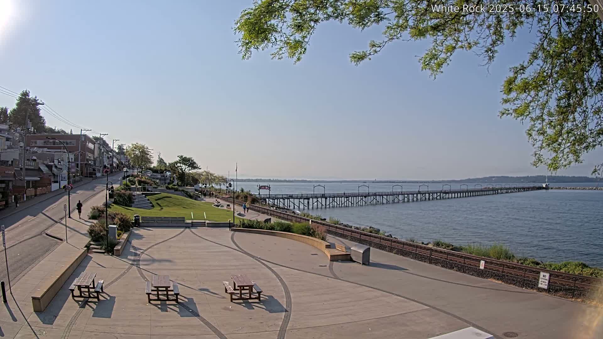 A paved waterfront area with picnic tables overlooks a long pier extending into a calm, blue bay under a clear, sunny sky.