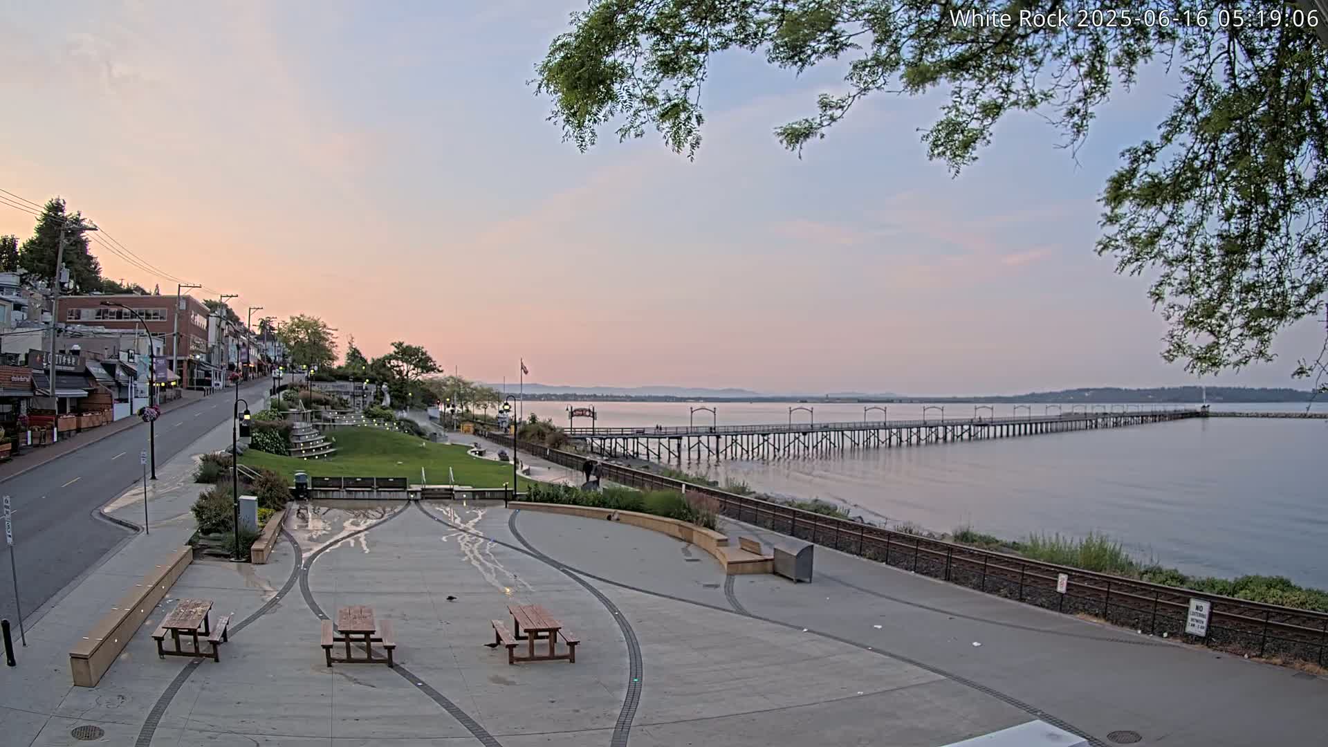 A waterfront town square with picnic tables overlooks a long pier extending into a calm sea under a pale sunset sky.