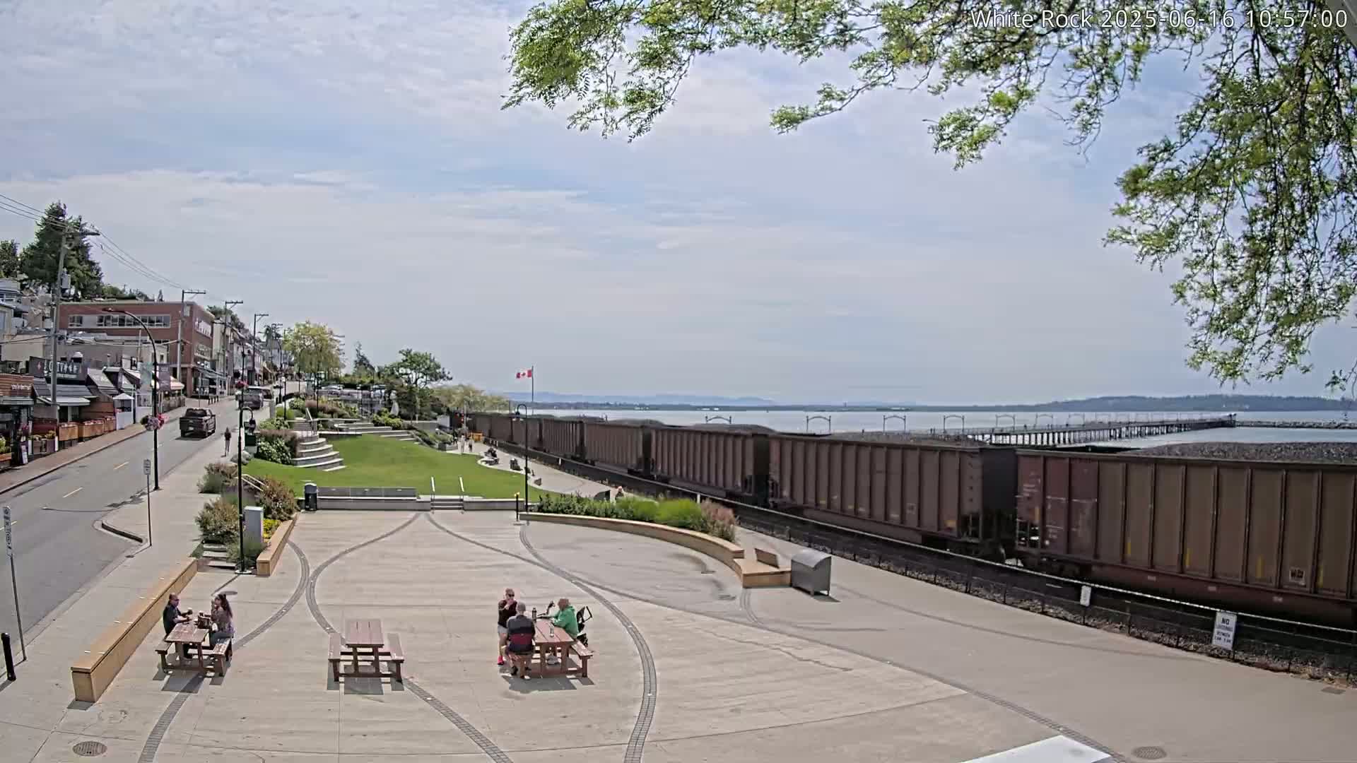 A partly sunny day reveals a waterfront town square with people sitting at picnic tables, a long freight train passing by, and a calm body of water extending to a distant shore.