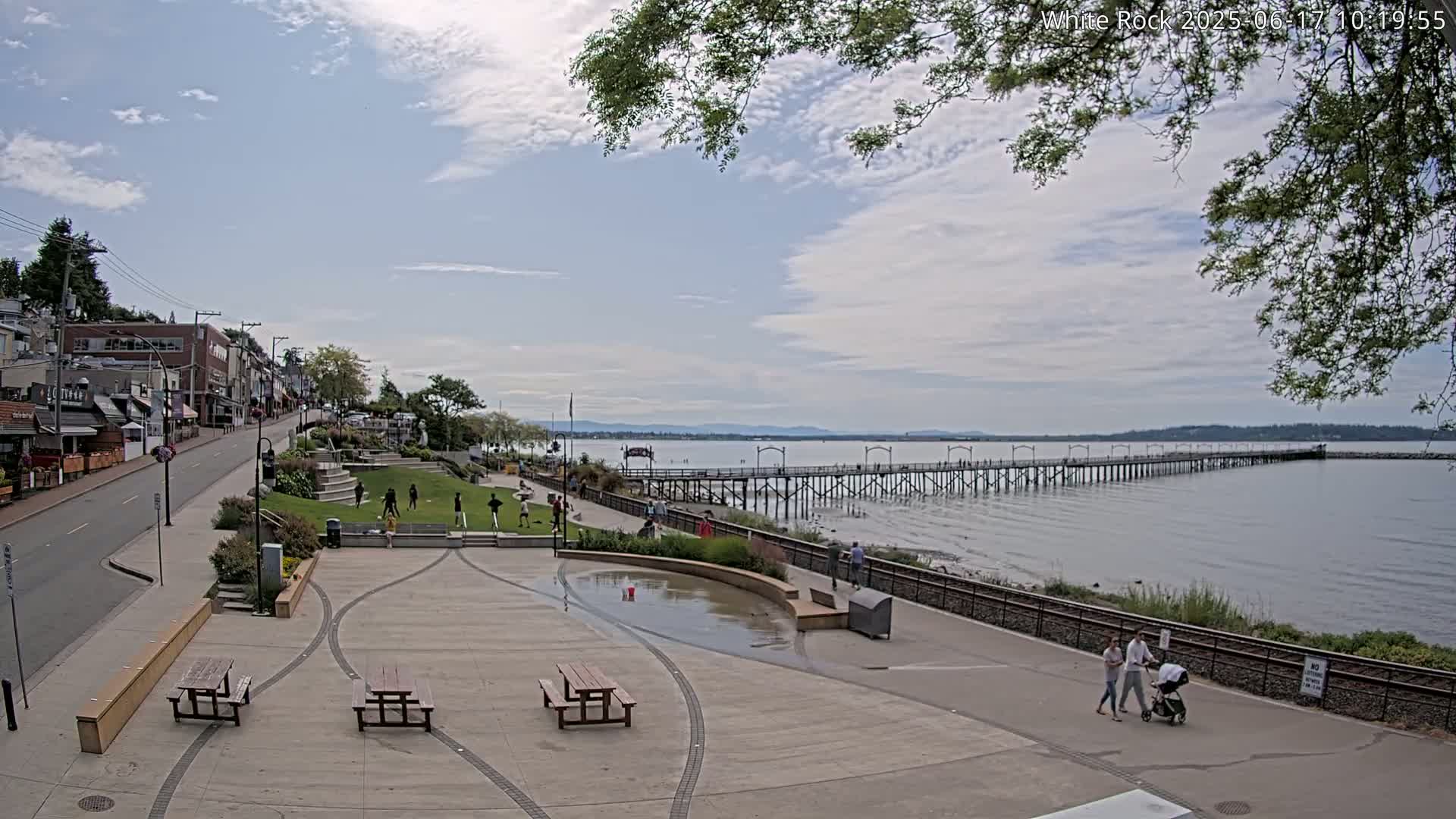 A waterfront town square with picnic tables and a long pier extending into a calm bay under a partly cloudy sky.