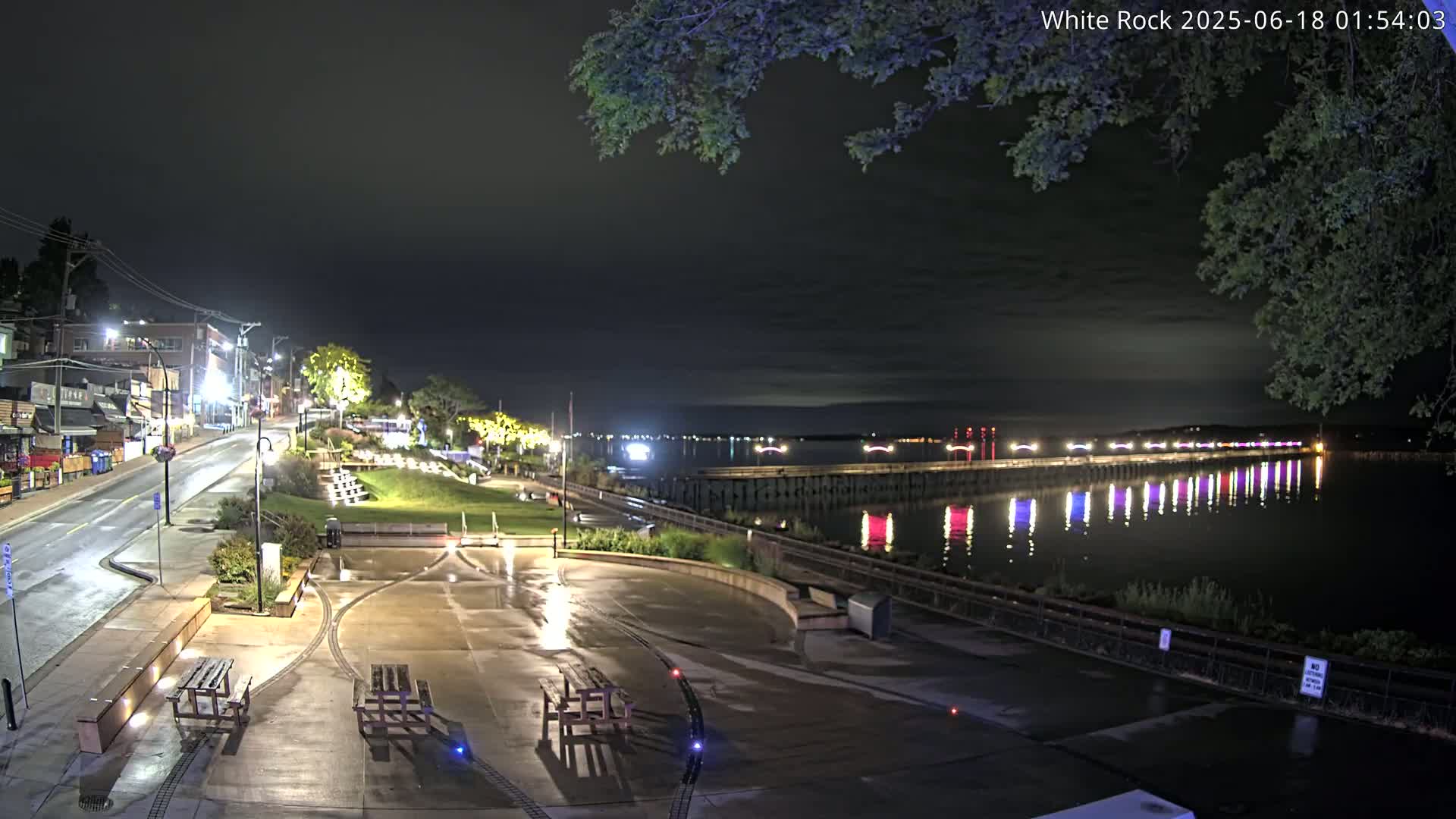 A nighttime view of a waterfront town with a wet paved plaza, picnic tables, and a long pier illuminated with colorful lights reflecting in the calm water under a dark, cloudy sky.