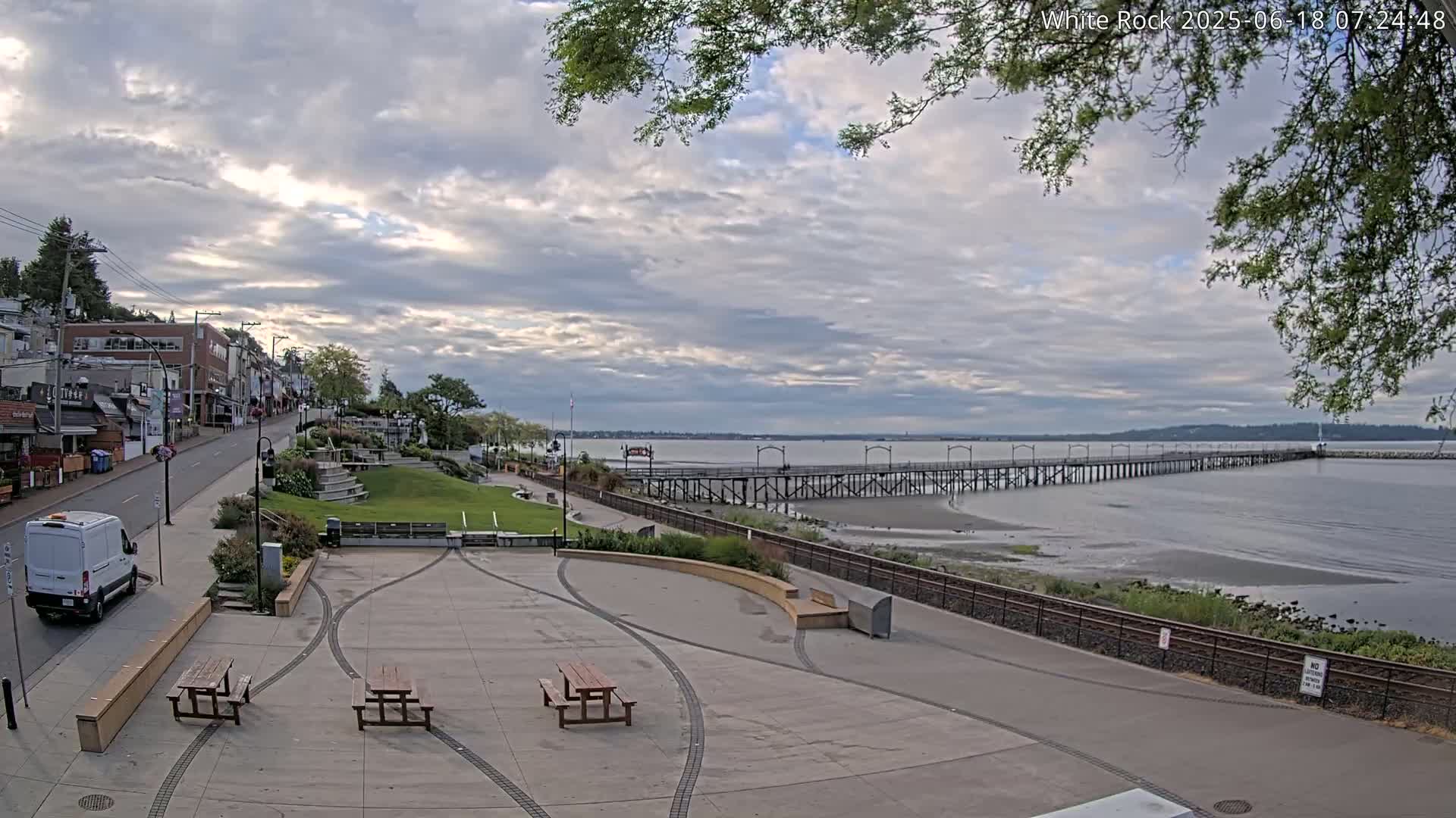 A paved plaza with picnic tables overlooks a long pier extending into a calm bay under a partly cloudy sky.
