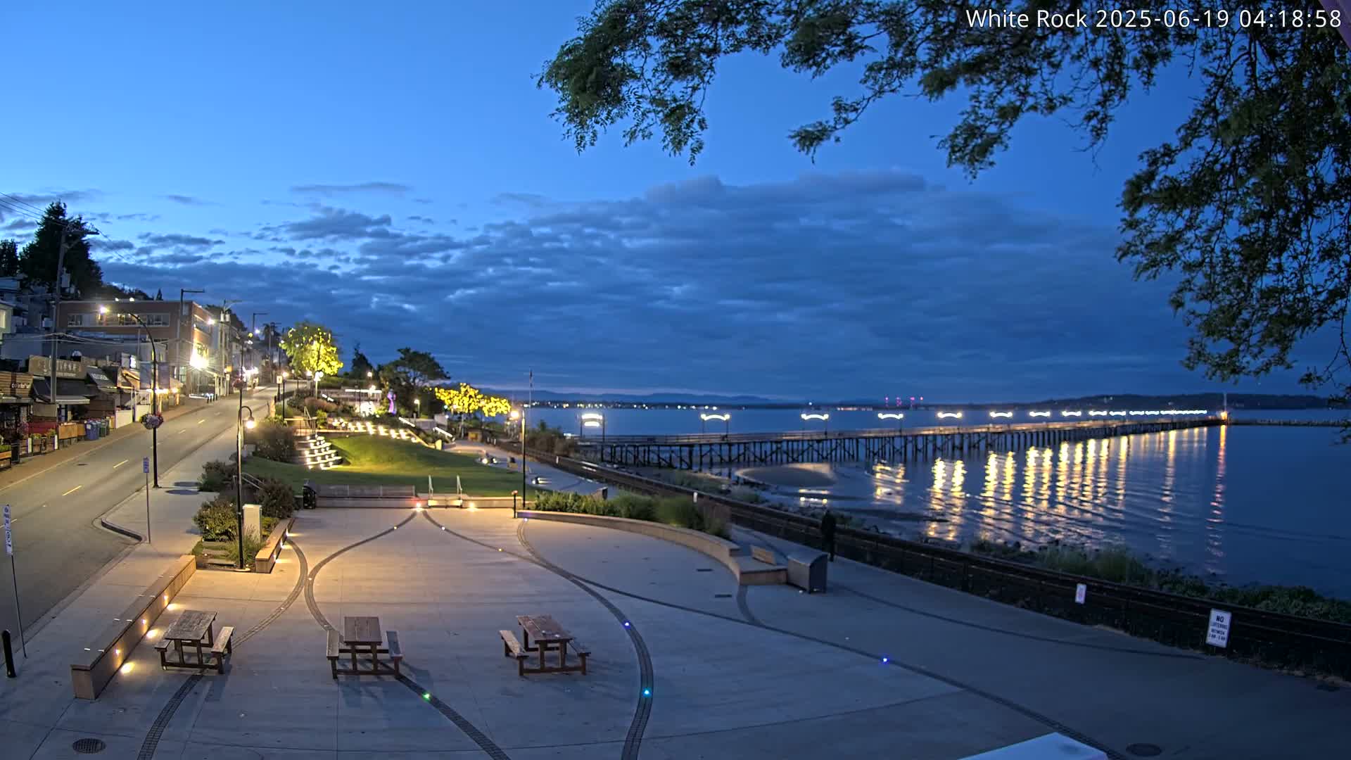 A waterfront town square at dusk, featuring picnic tables, a paved walkway, and a long pier extending into a calm body of water under a partly cloudy sky.