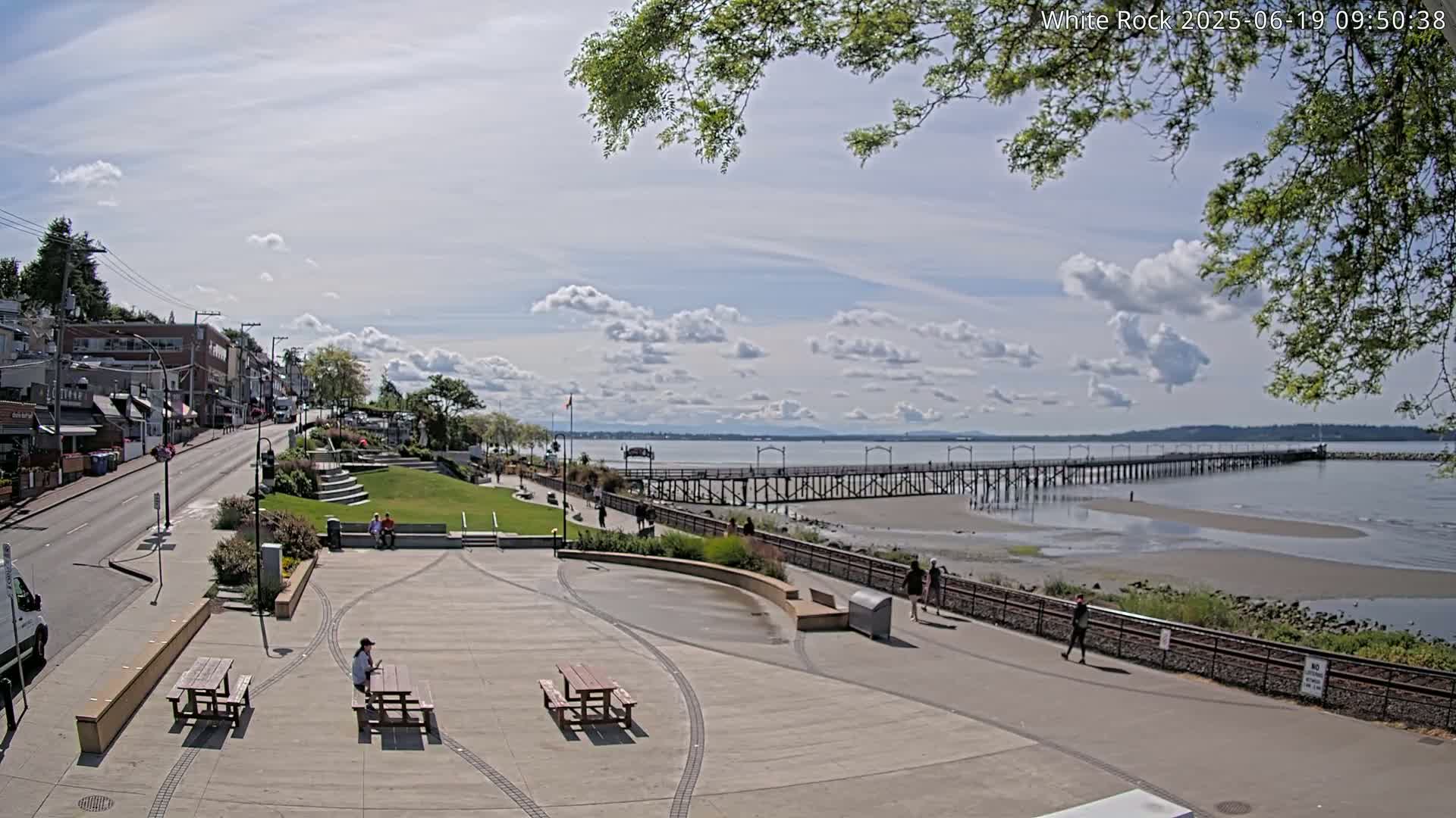 A partly sunny day shows a waterfront town square with picnic tables and a long pier extending into a calm bay under a mostly cloudy sky.
