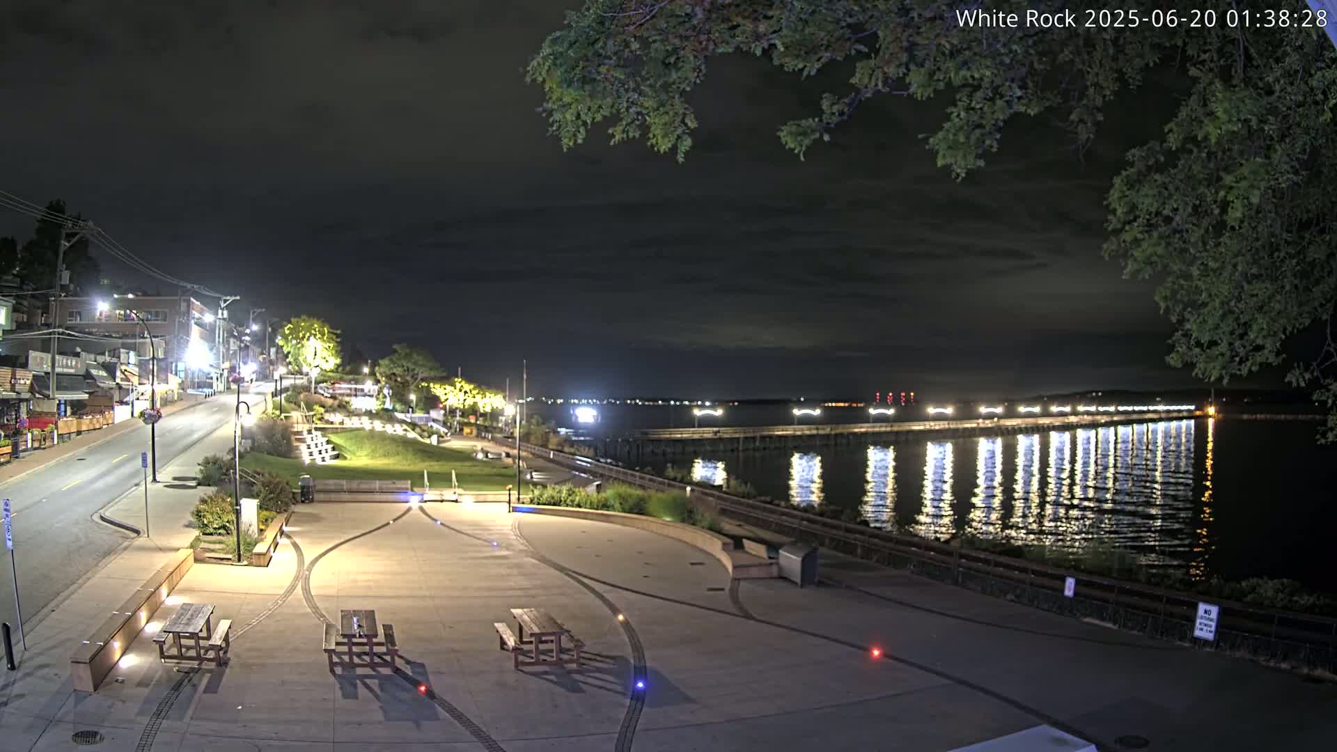 A nighttime view of a waterfront park with picnic tables, a paved walkway, and a long pier extending into calm water under a partly cloudy sky.