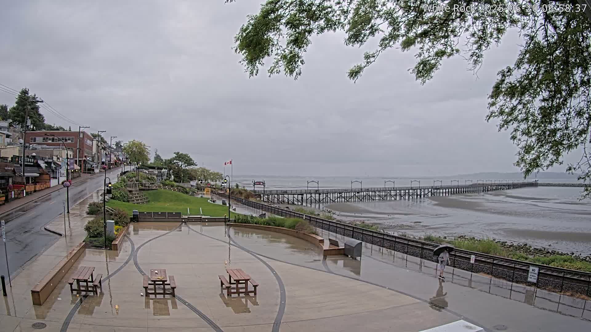 A wet plaza with picnic tables overlooks a long pier extending into a calm, gray sea under an overcast sky.