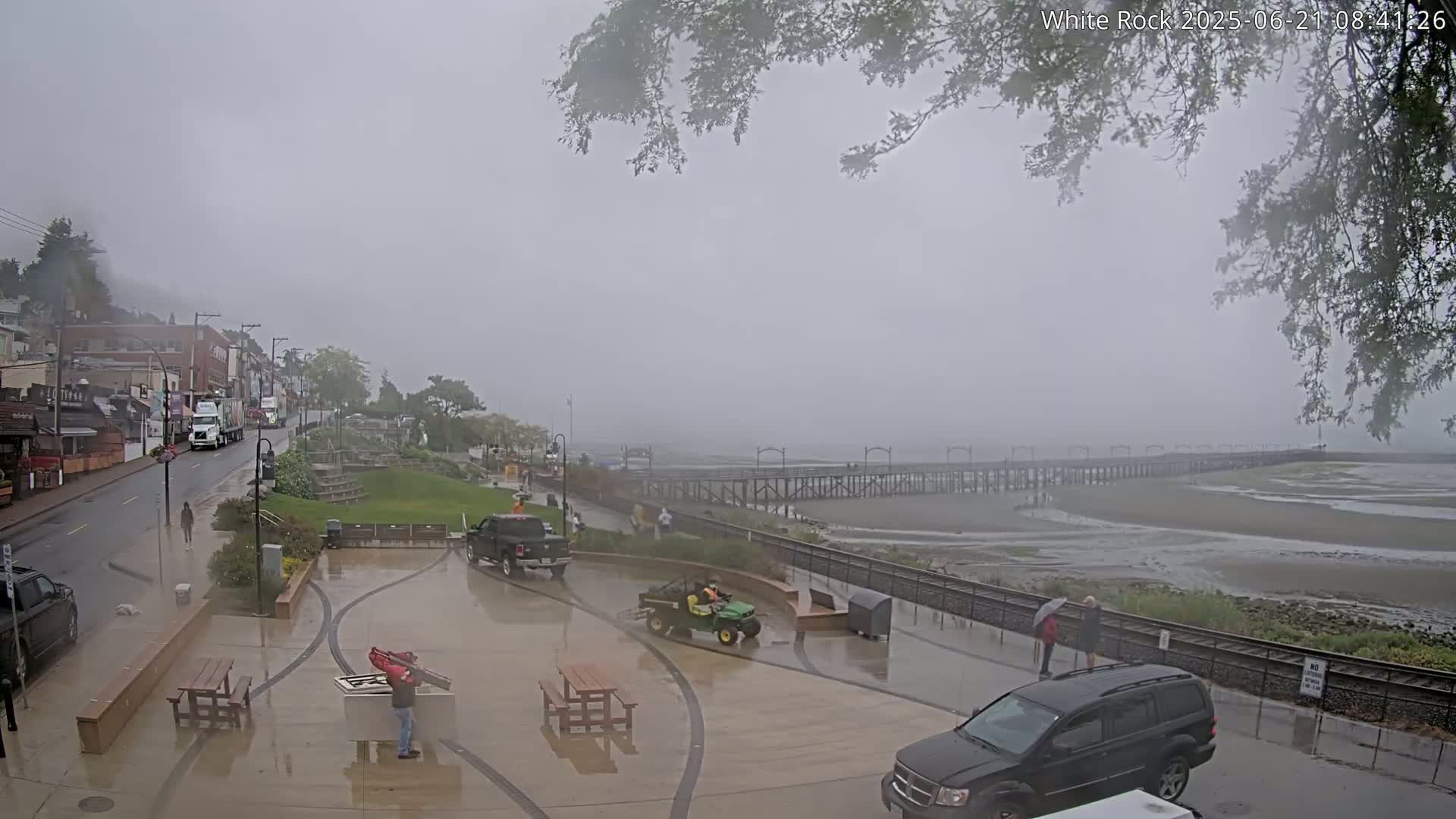 A wet, foggy seaside town scene shows a long pier extending into shallow, murky water, with vehicles and people visible along a paved waterfront area and a street lined with buildings.