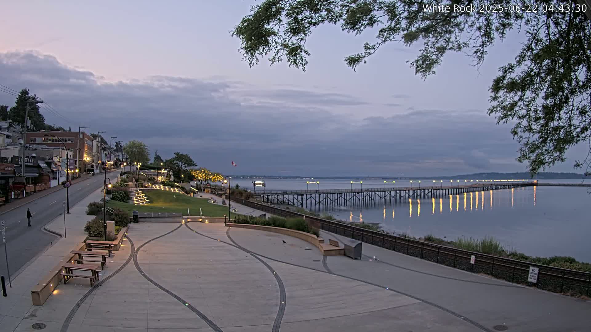A paved waterfront plaza with benches and a long pier extending into a calm, twilight ocean under a cloudy sky.
