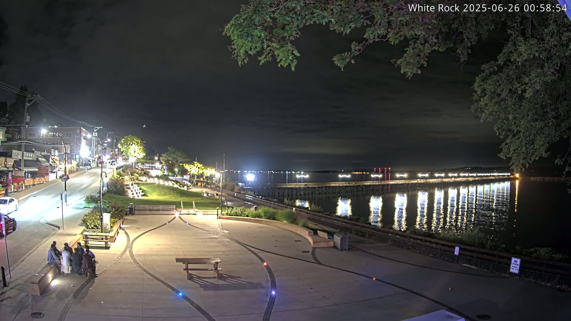 A nighttime view of a waterfront plaza with a long pier extending into a calm body of water under a dark, cloudy sky, with a street and buildings visible in the background.