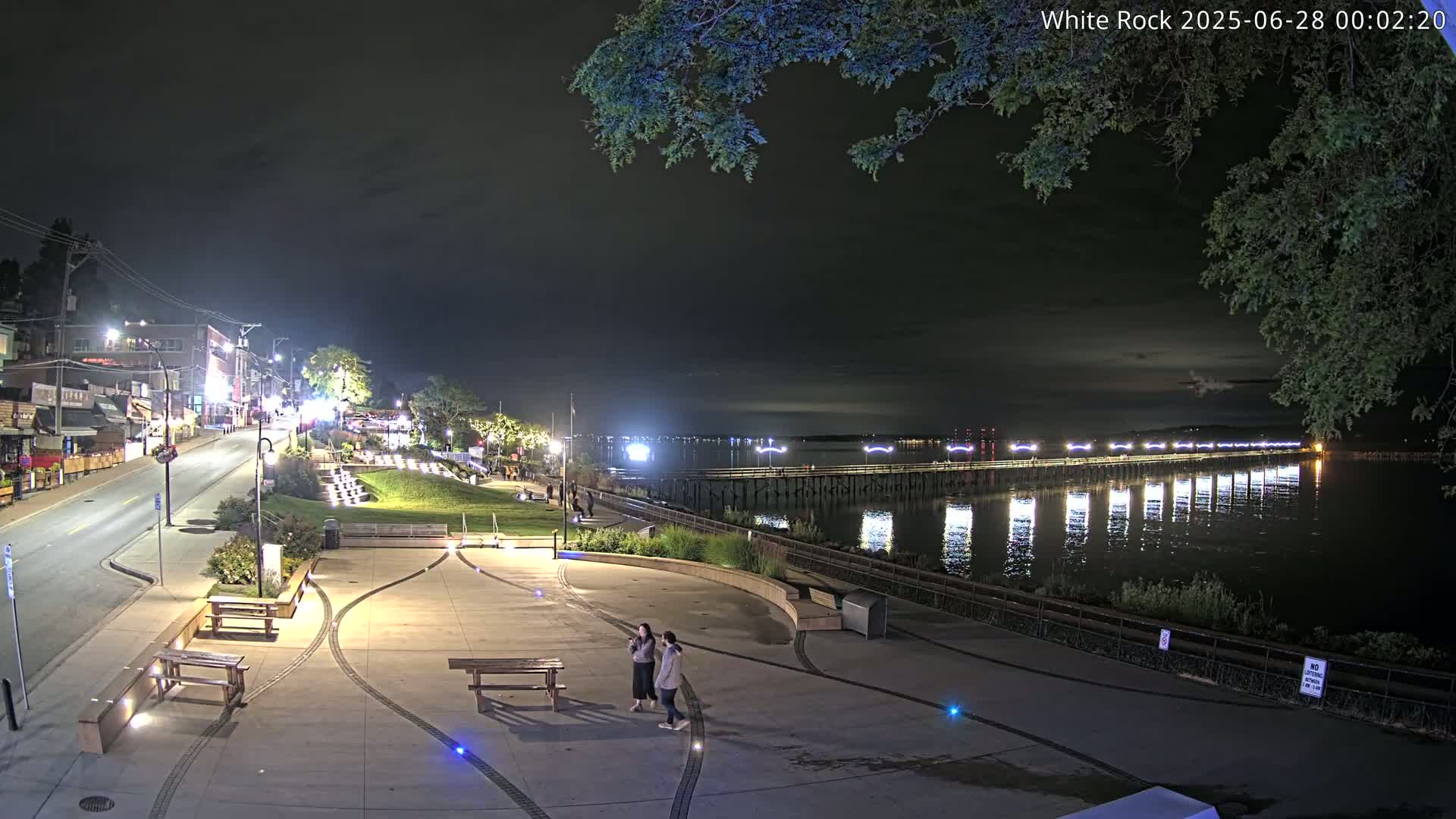 A nighttime view of a waterfront town with a long pier extending into calm water, two people standing in a plaza, and streetlights illuminating the scene.