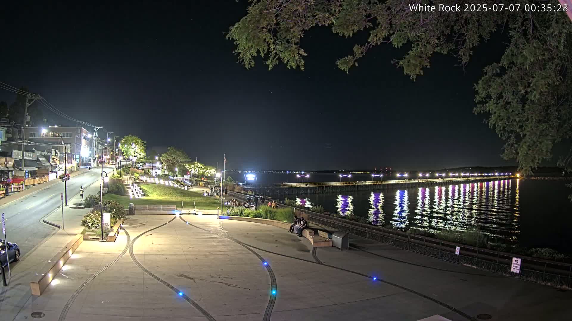 A nighttime view of a waterfront plaza with a long pier extending into a calm body of water, illuminated by lights reflecting on the surface.