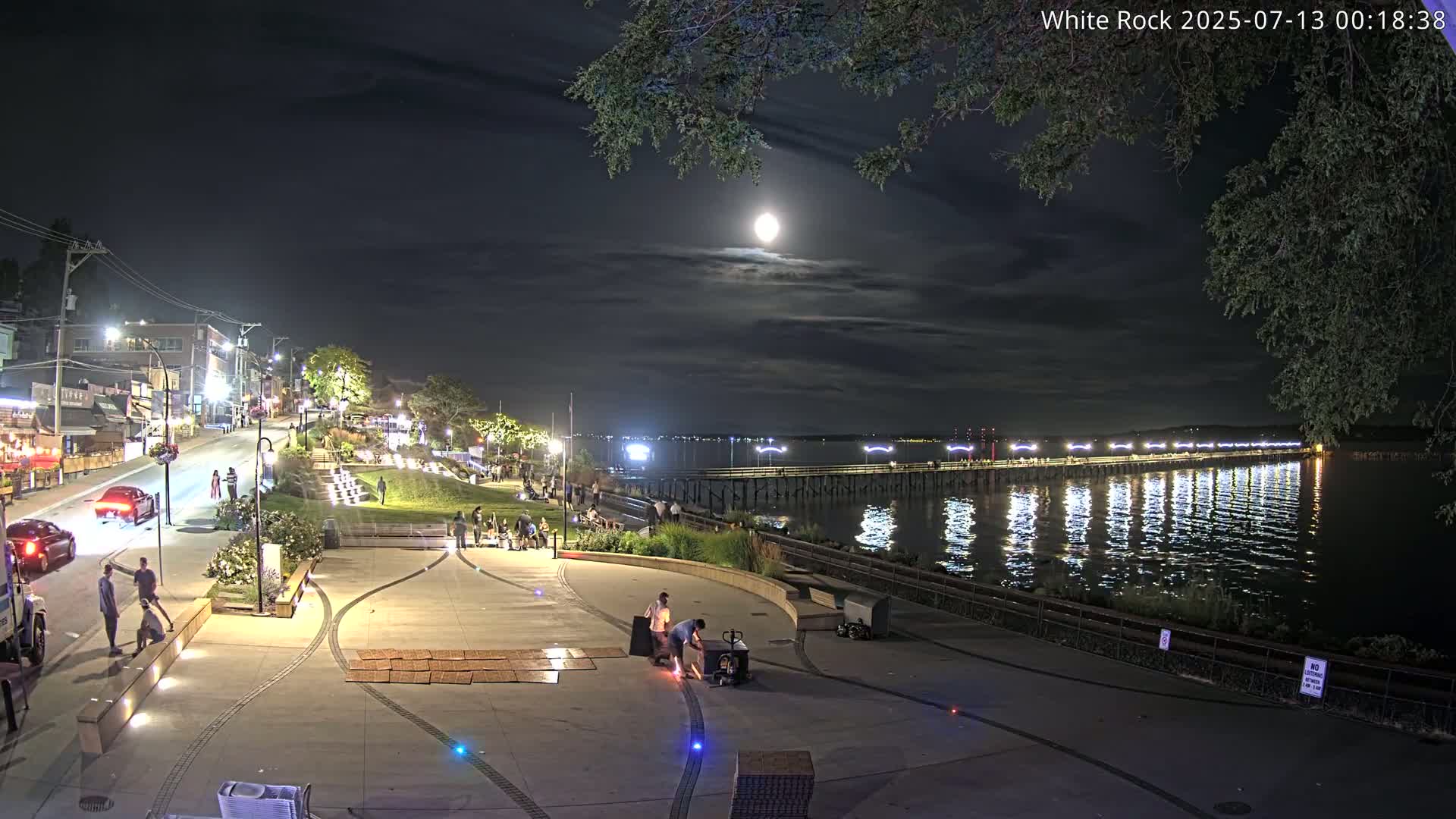A nighttime scene shows a waterfront plaza with people gathered, cars parked along a street, and a brightly lit pier extending into calm water under a full moon and partly cloudy sky.