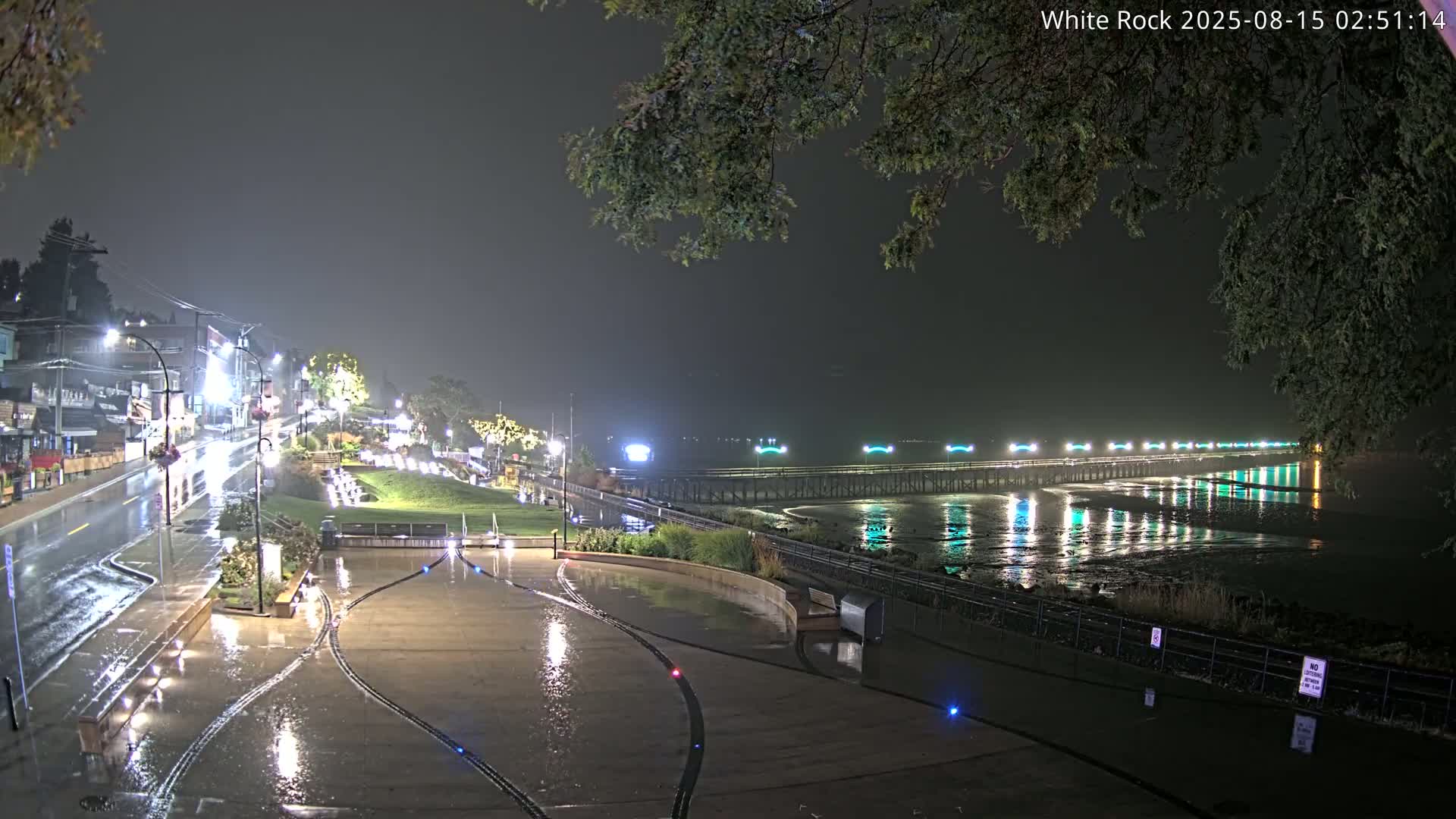 A brightly lit pier extends into a calm body of water at night, its lights reflecting on the surface, with a paved plaza and a street lined with buildings visible in the foreground.