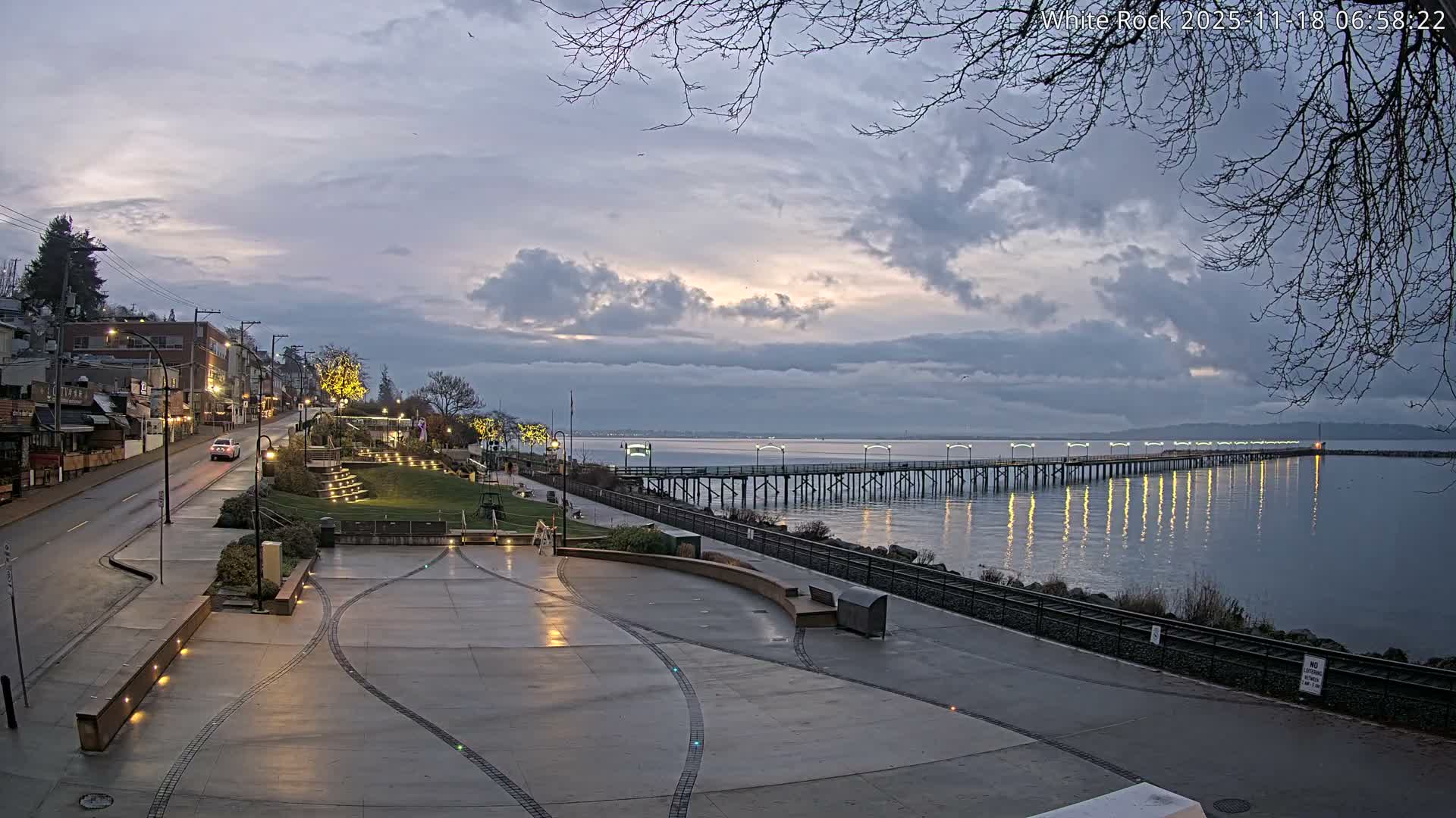 A nighttime view depicts a waterfront town with a long, illuminated pier extending into dark, reflective water, while wet pavement and roads, glimmering with reflections from abundant lights, indicate recent rain under an overcast sky.