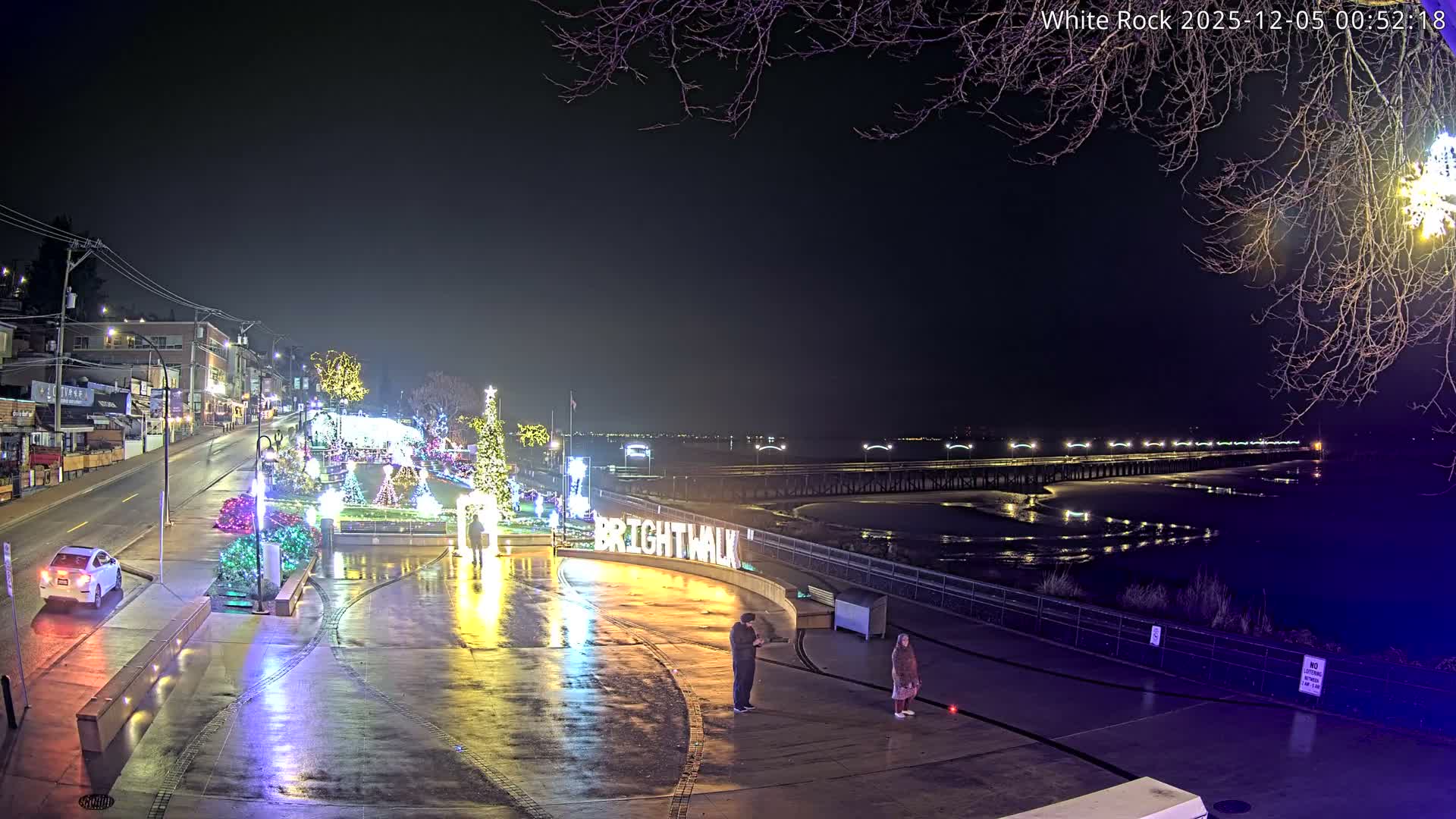 On a damp night, a brightly illuminated waterfront promenade is adorned with colorful festive lights and Christmas decorations, featuring people, a wet street with a single car, and a long pier stretching into the dark body of water.