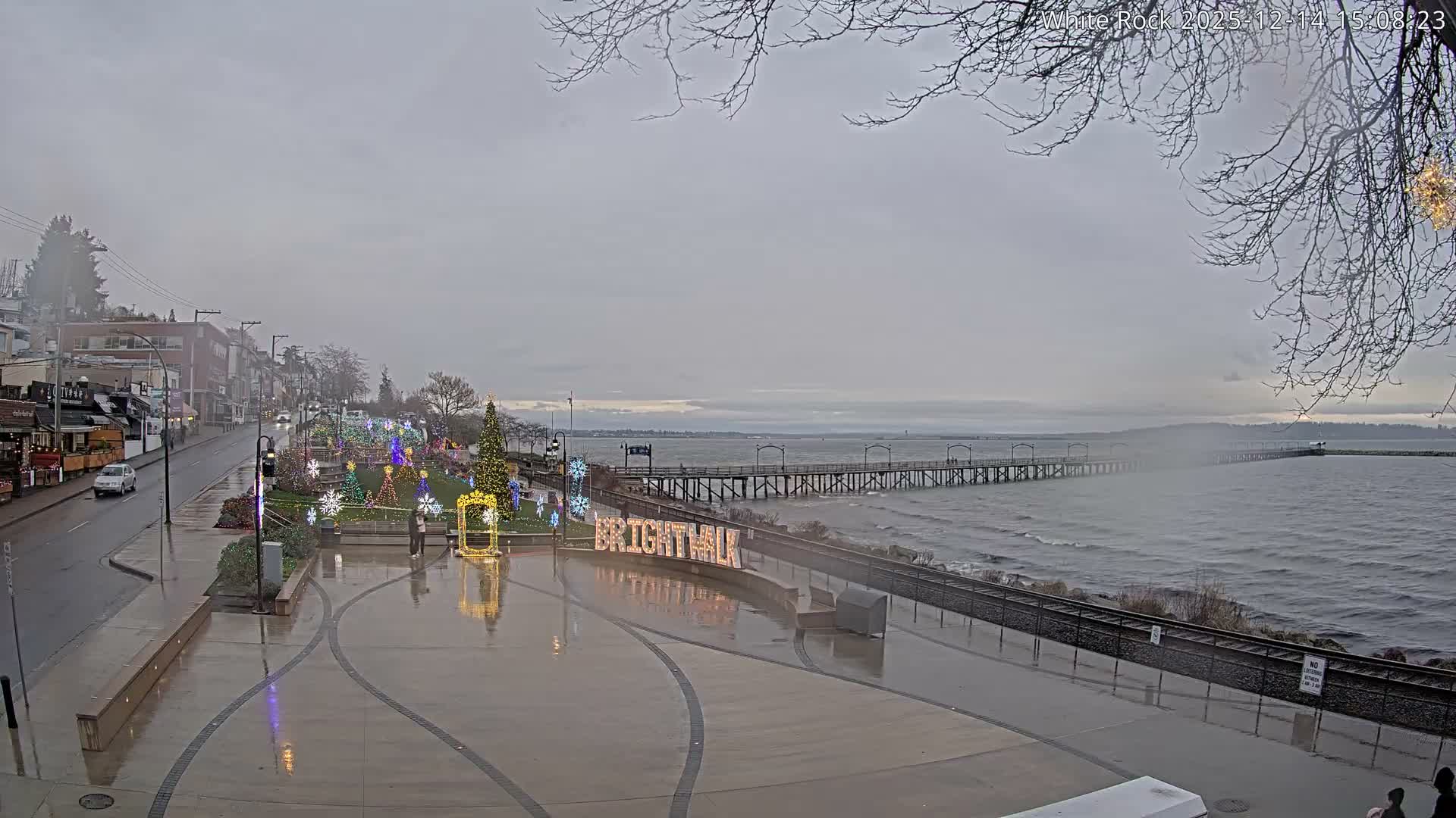 On a damp night, a brightly illuminated waterfront promenade is adorned with colorful festive lights and Christmas decorations, featuring people, a wet street with a single car, and a long pier stretching into the dark body of water.