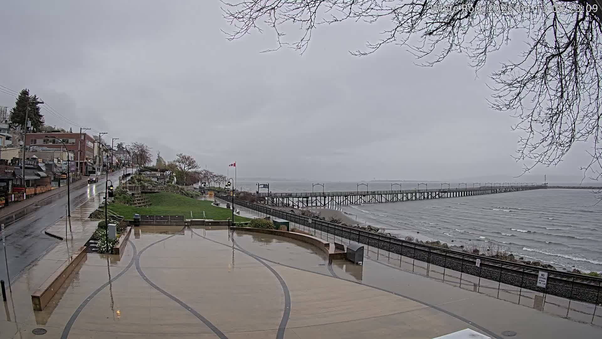 On a damp night, a brightly illuminated waterfront promenade is adorned with colorful festive lights and Christmas decorations, featuring people, a wet street with a single car, and a long pier stretching into the dark body of water.