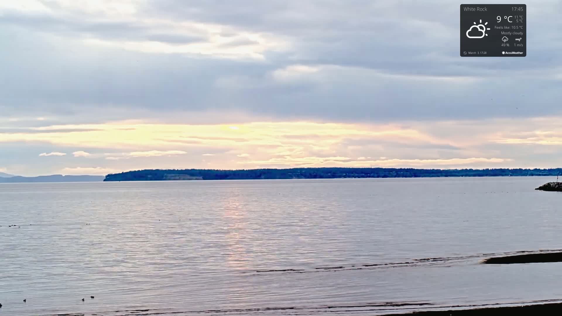 A mostly cloudy sunset over a calm body of water with a distant shoreline.