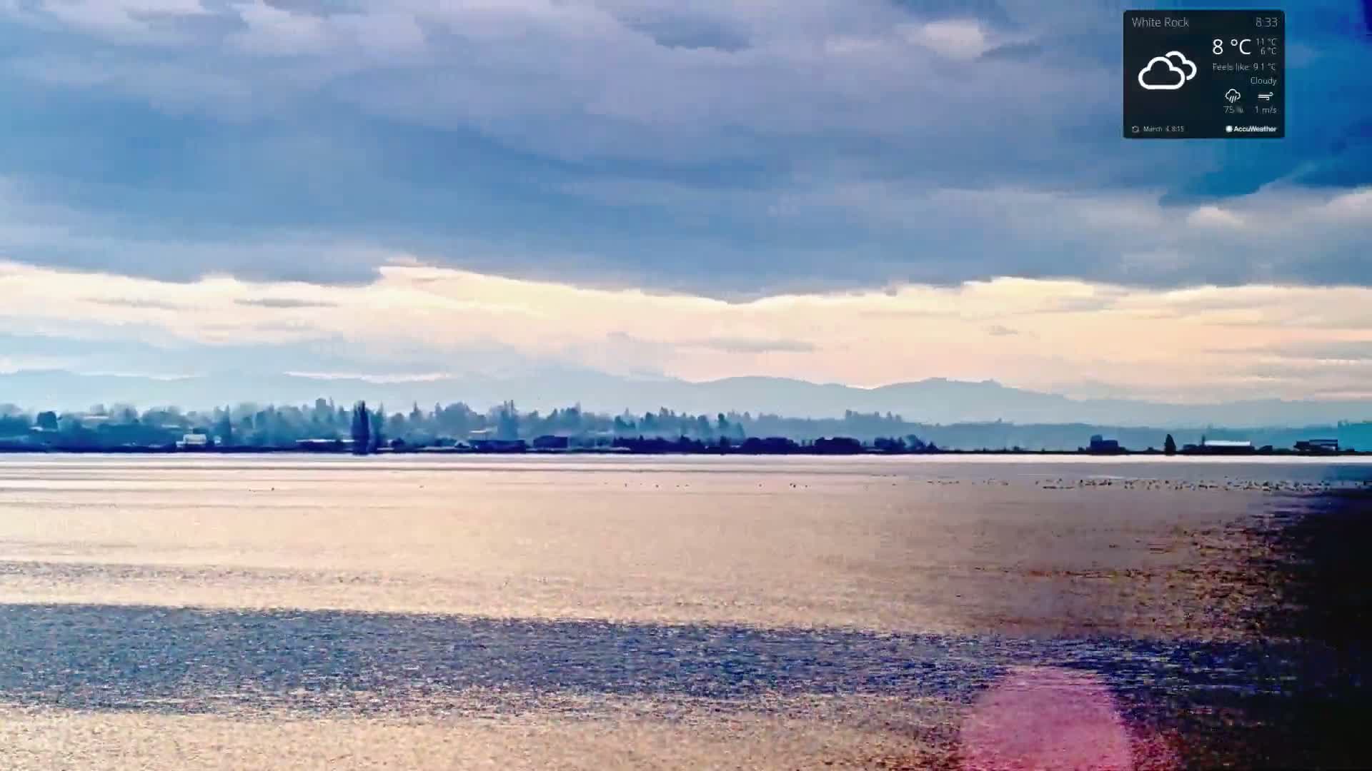 A calm body of water reflects a cloudy sky and distant, hazy mountains under a partly cloudy sky.