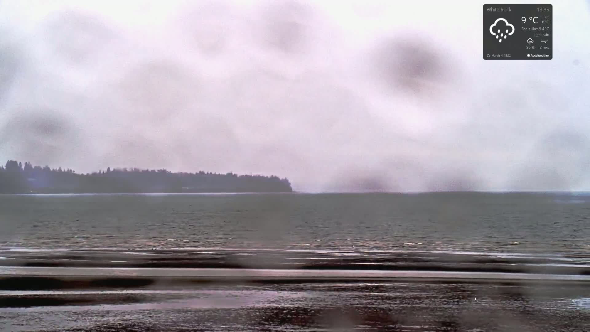 A dark, flat shoreline stretches across a calm body of water under a gray, overcast sky with light rain.