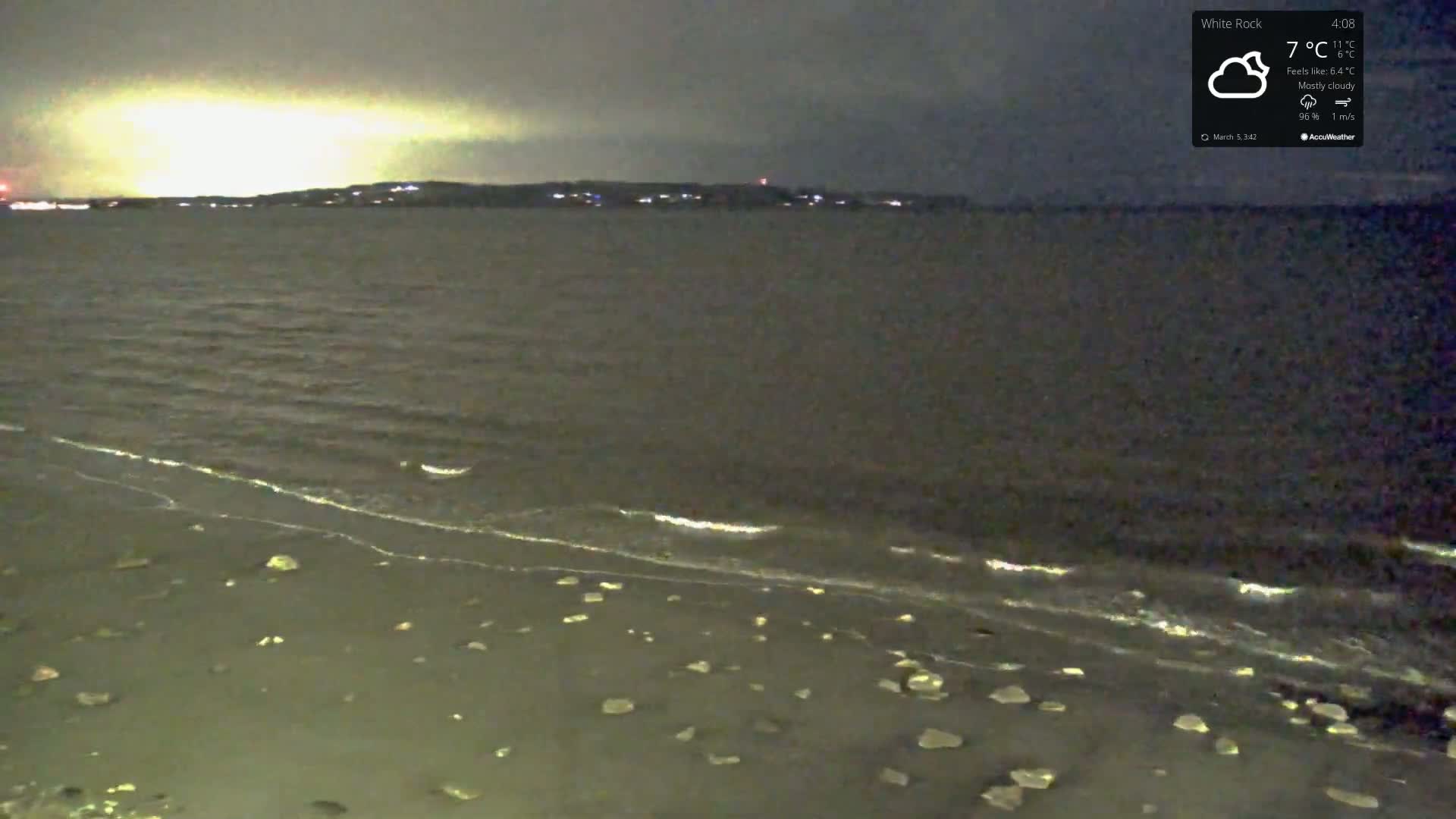 A mostly cloudy night scene shows a dark body of water lapping gently at a rocky shoreline under a pale, yellowish sky.