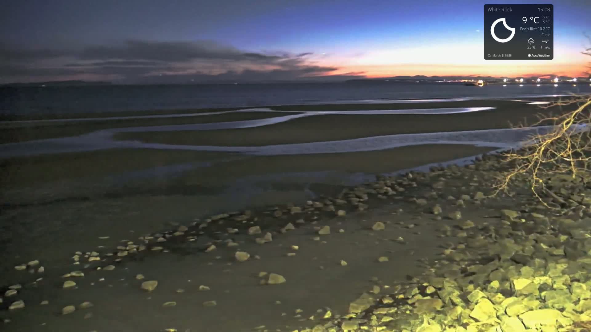 A rocky shoreline at dusk under a clear sky shows a calm body of water with exposed mudflats at low tide.