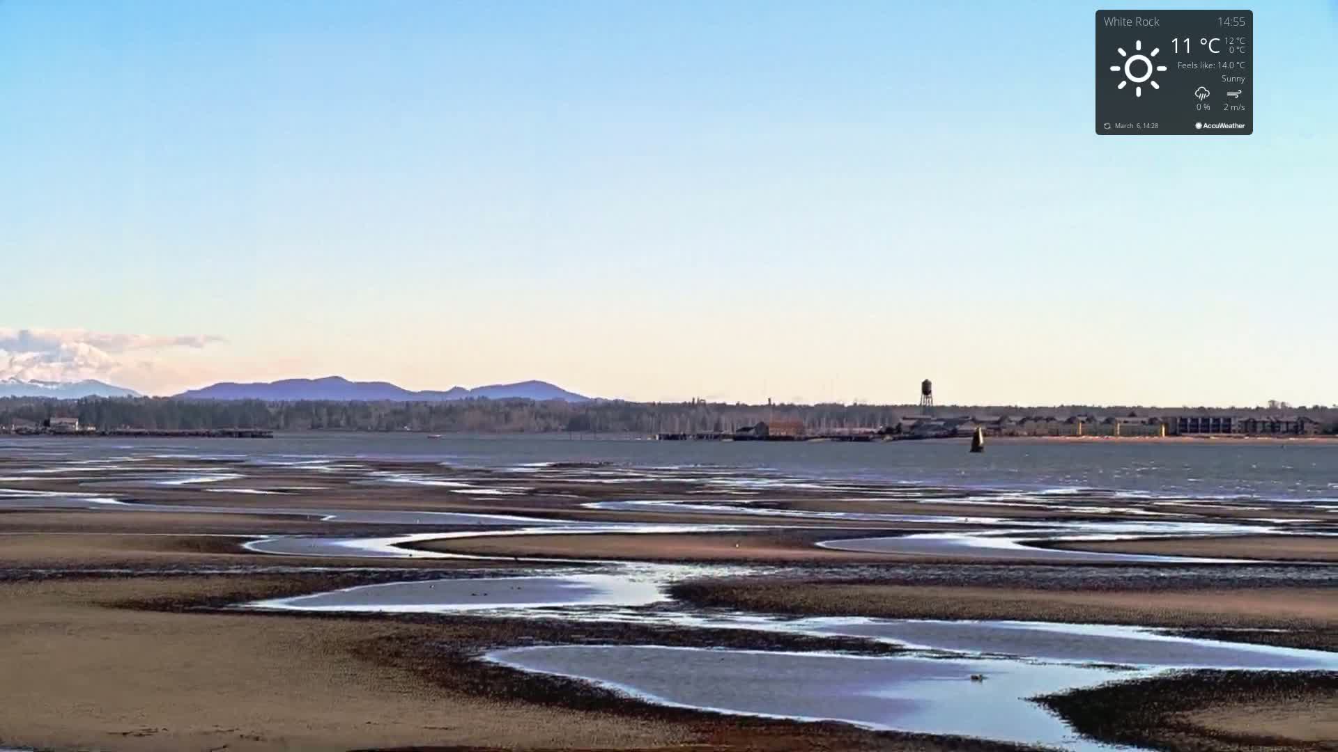 A sunny day reveals a wide expanse of tidal flats with winding channels of water, backed by a calm body of water, distant mountains, and a shoreline with buildings.