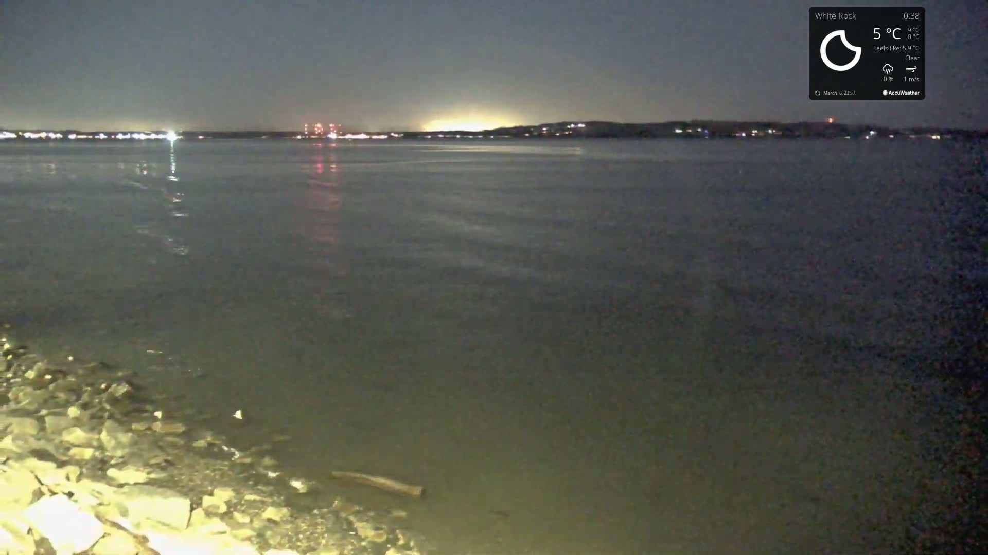 A nighttime view of a calm body of water under a clear sky, with distant lights visible on the opposite shore and a rocky shoreline in the foreground.