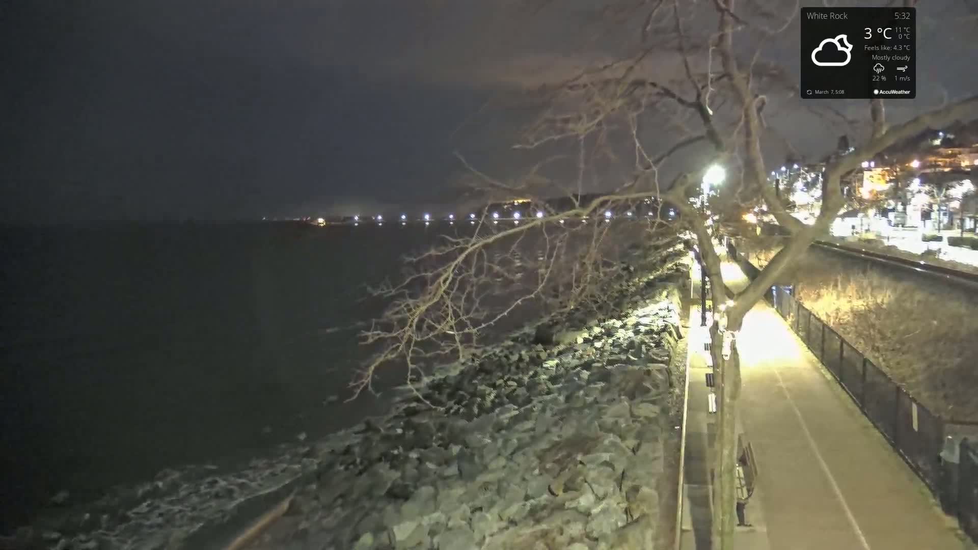 A nighttime view of a rocky shoreline with a paved path illuminated by lights under a mostly cloudy sky.
