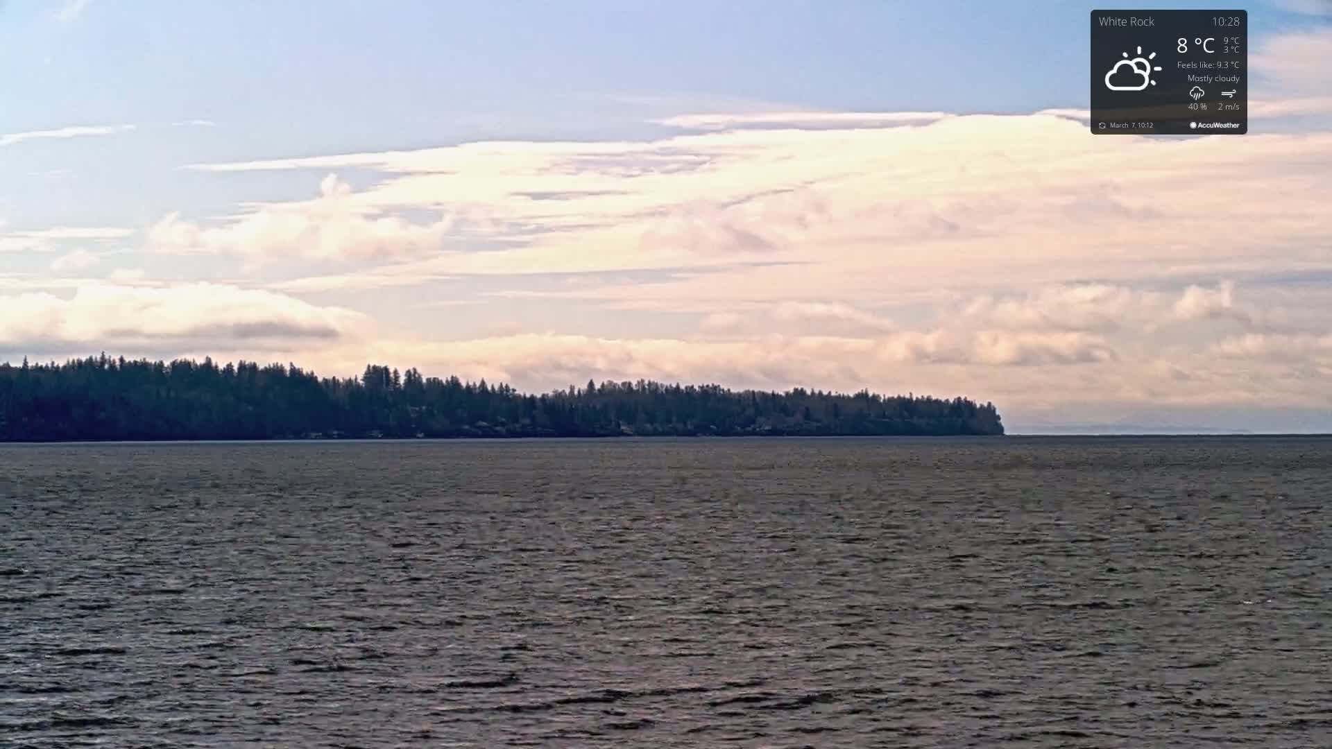 A mostly cloudy sky over a calm body of water with a dark, tree-covered shoreline in the distance.