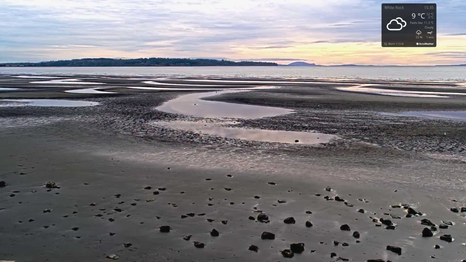 A cloudy, 9°C day reveals a wide expanse of tidal flats with numerous shallow pools of water at low tide, stretching towards a distant shoreline under a pale sky.
