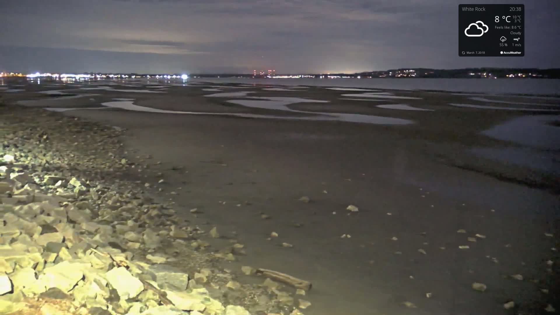 A rocky shoreline at night under cloudy skies shows a dark, tidal flat with scattered pools of water and distant city lights.