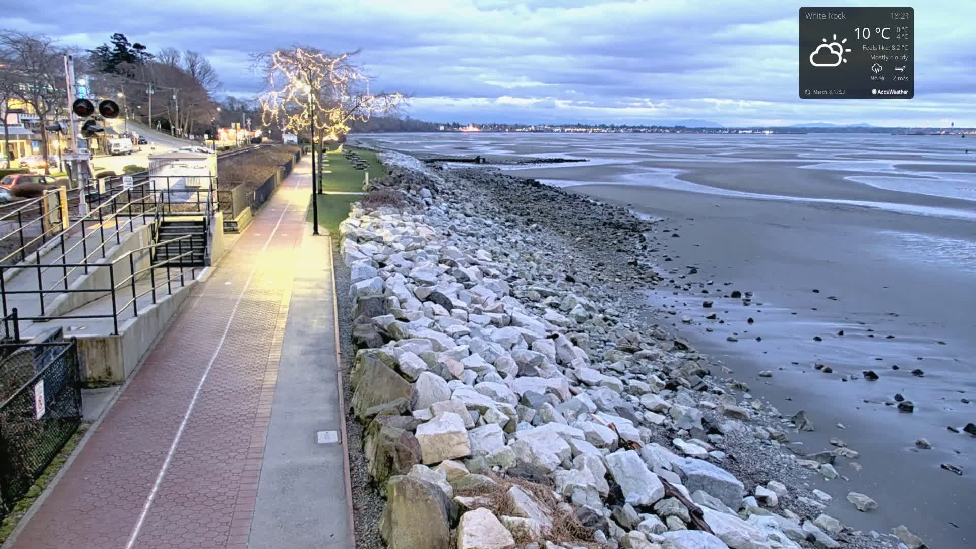 A paved walkway next to a rocky shoreline overlooks a calm, mostly cloudy, gray beach at twilight.