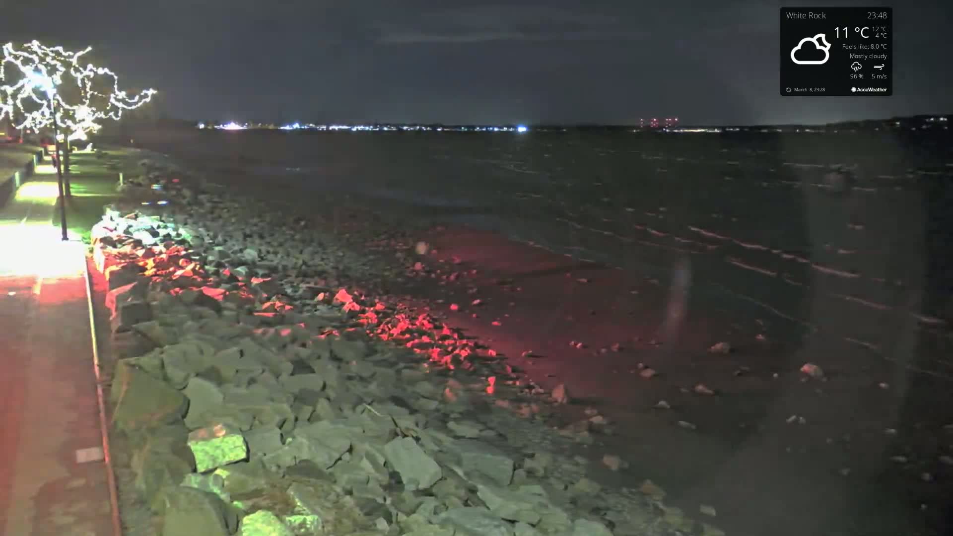A rocky shoreline at night under mostly cloudy skies, illuminated by lights along a paved walkway and faint city lights across the water.